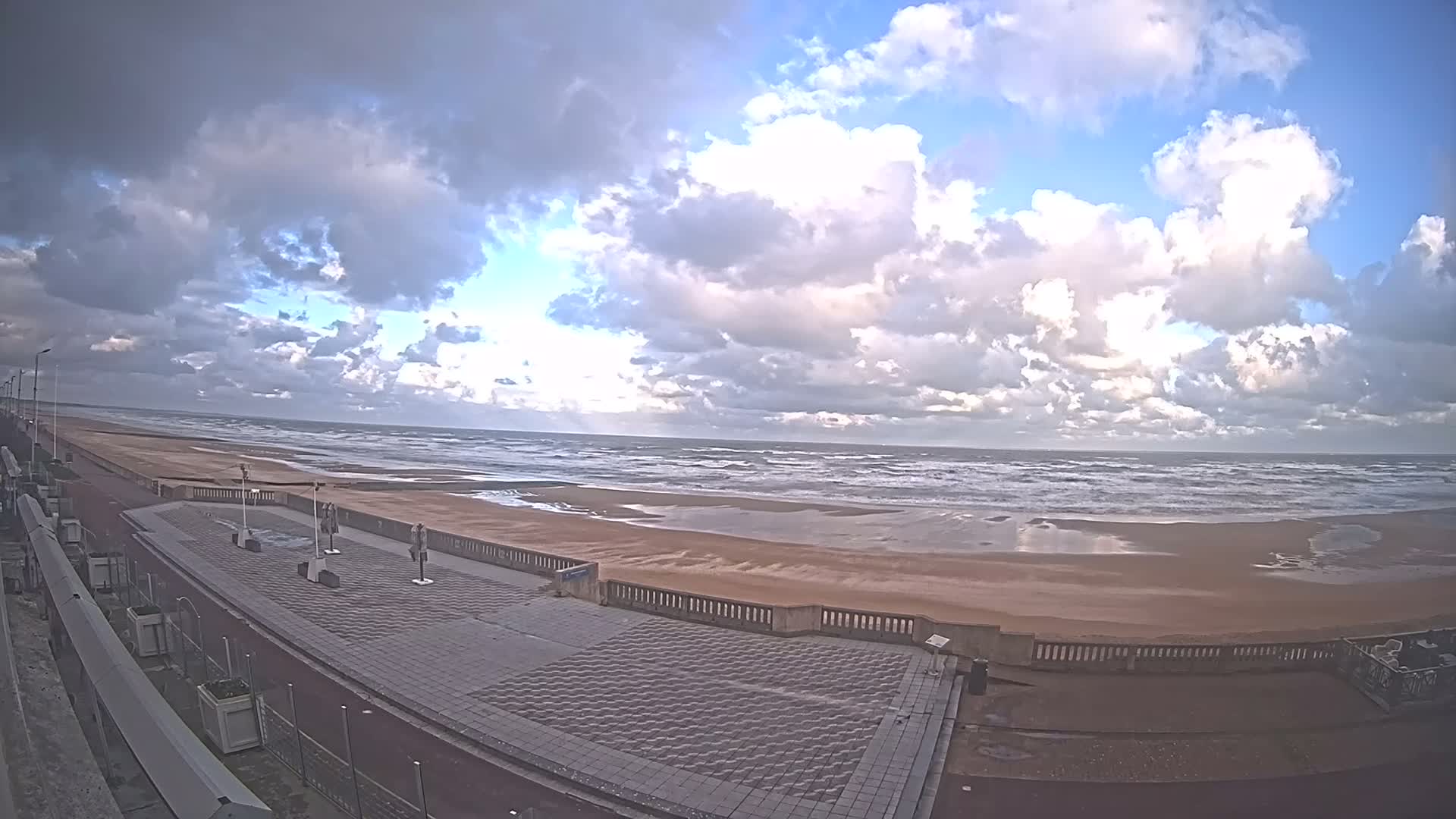 A wide sandy beach with active waves meets a paved promenade under a partly cloudy sky, with bright blue patches visible between large, fluffy clouds.