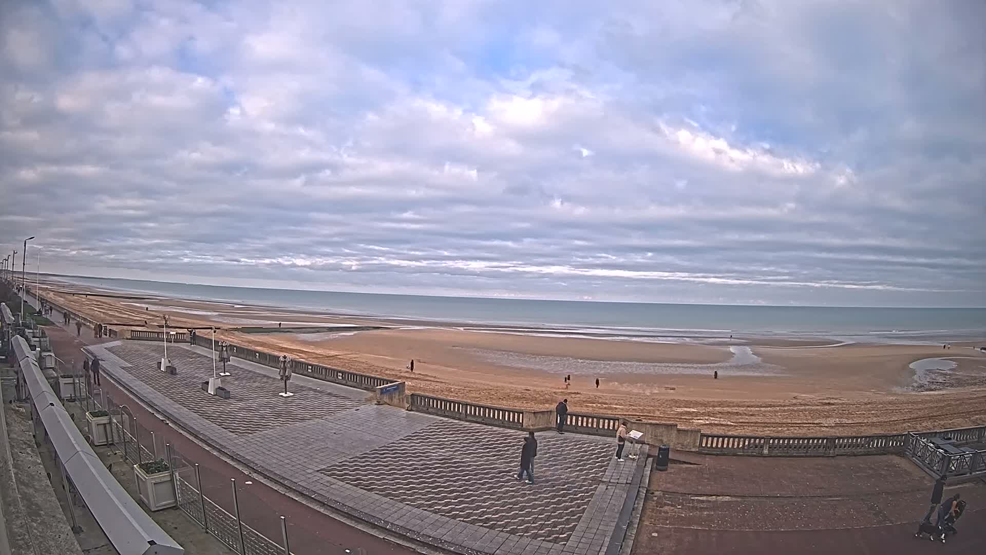 A wide sandy beach with active waves meets a paved promenade under a partly cloudy sky, with bright blue patches visible between large, fluffy clouds.