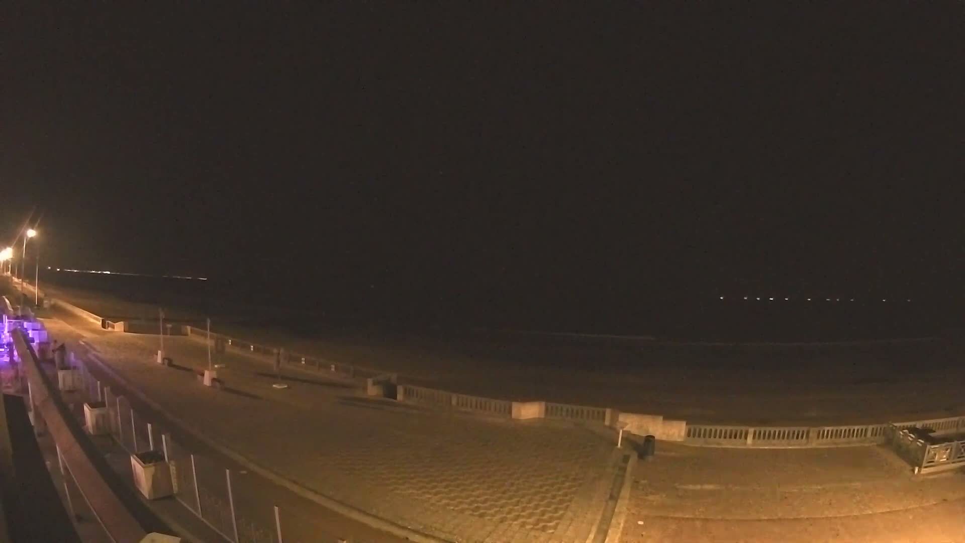 A wide sandy beach with active waves meets a paved promenade under a partly cloudy sky, with bright blue patches visible between large, fluffy clouds.