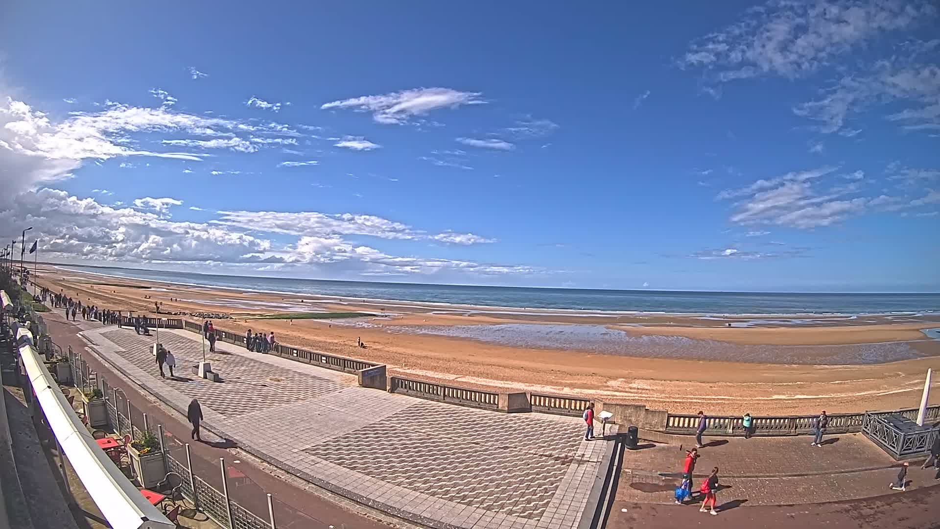 A wide sandy beach with active waves meets a paved promenade under a partly cloudy sky, with bright blue patches visible between large, fluffy clouds.