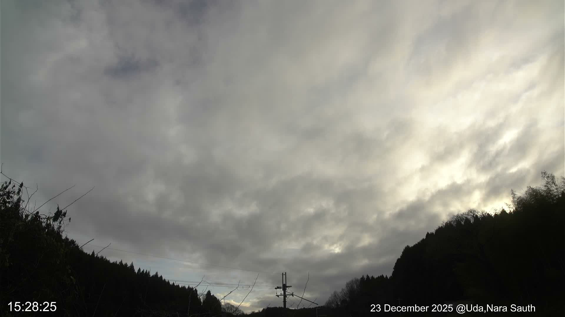 An expansive view reveals a predominantly overcast sky with dense grey and brighter white clouds, looming above dark, silhouetted hills and trees, with a visible utility pole.