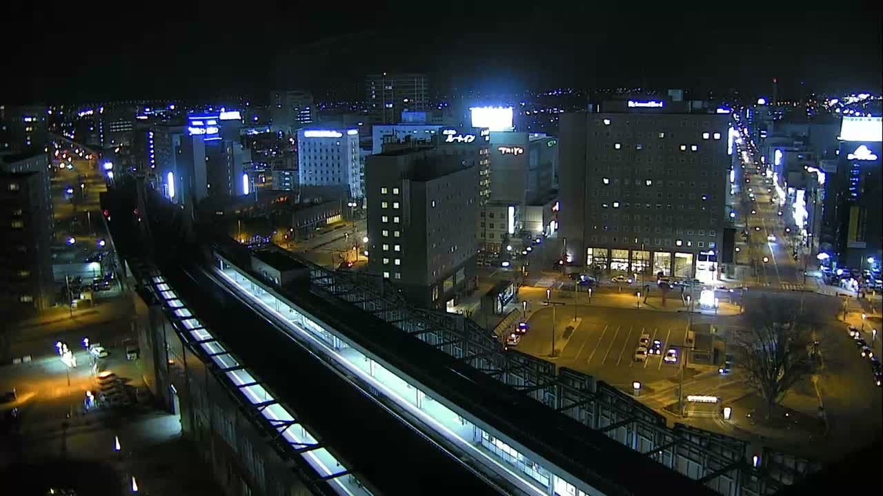An aerial view of a snowy city at night reveals a network of illuminated buildings and streets under a clear, dark sky, with a dark elevated structure curving through the foreground.