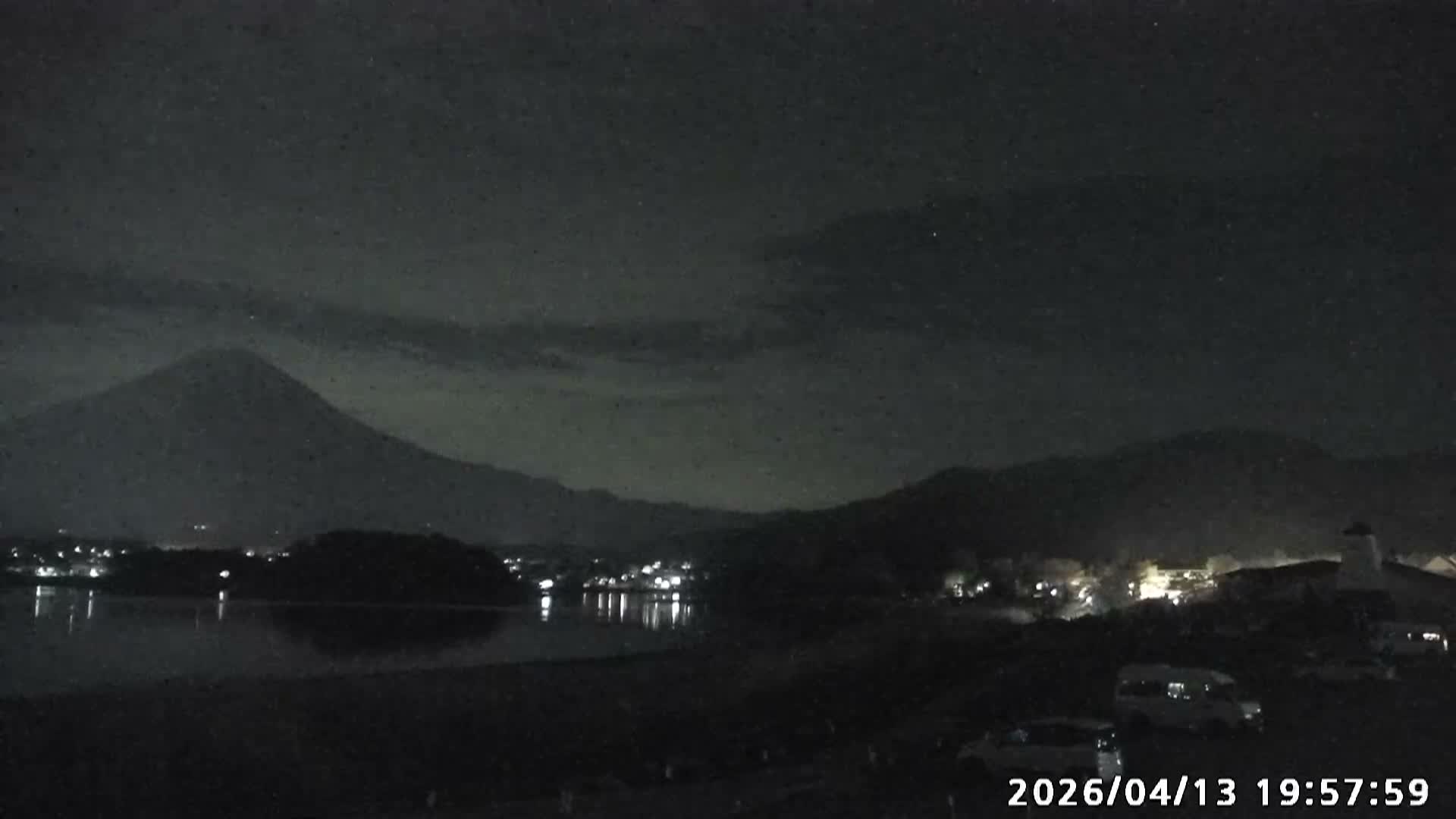 A nighttime view across a body of water reveals the distant lights of a town reflecting on its surface under a dark, cloudy sky.