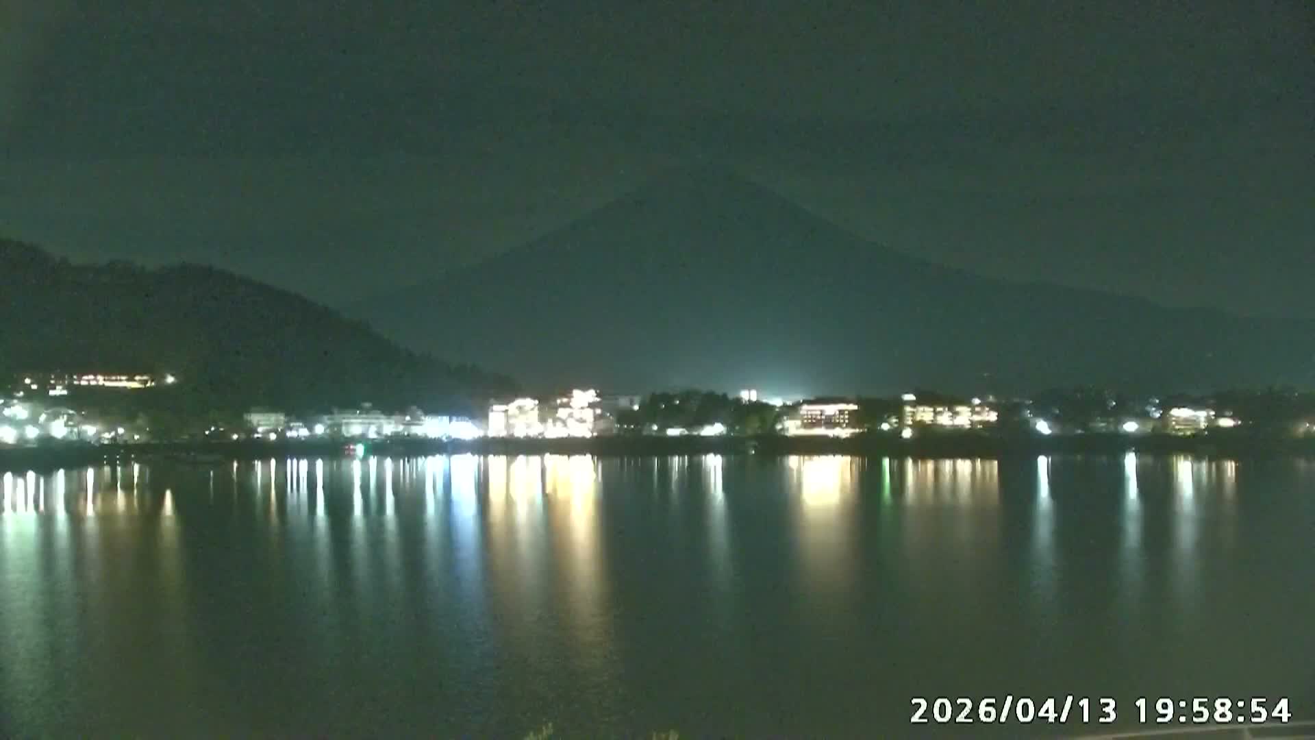 A clear winter morning illuminates the snow-capped peak of Mount Fuji with golden light, overlooking a partially frozen lake and a town nestled along its dark, forested shore.