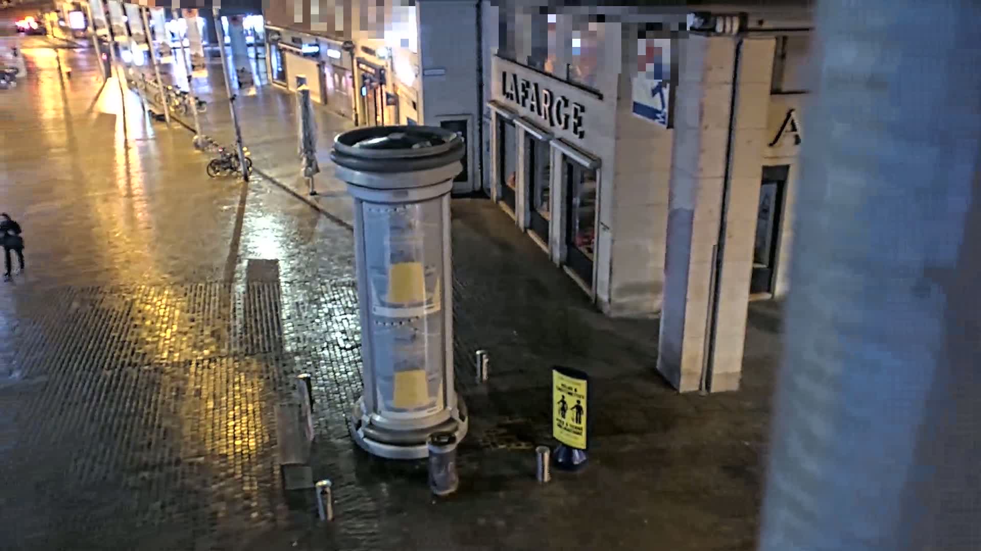 A wet, nighttime city street scene shows a person walking past a cylindrical advertisement display near a building.