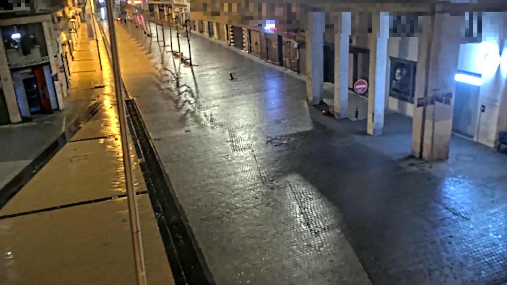 A wet, empty street at night is lit by streetlights, showing the reflections of light on the pavement.