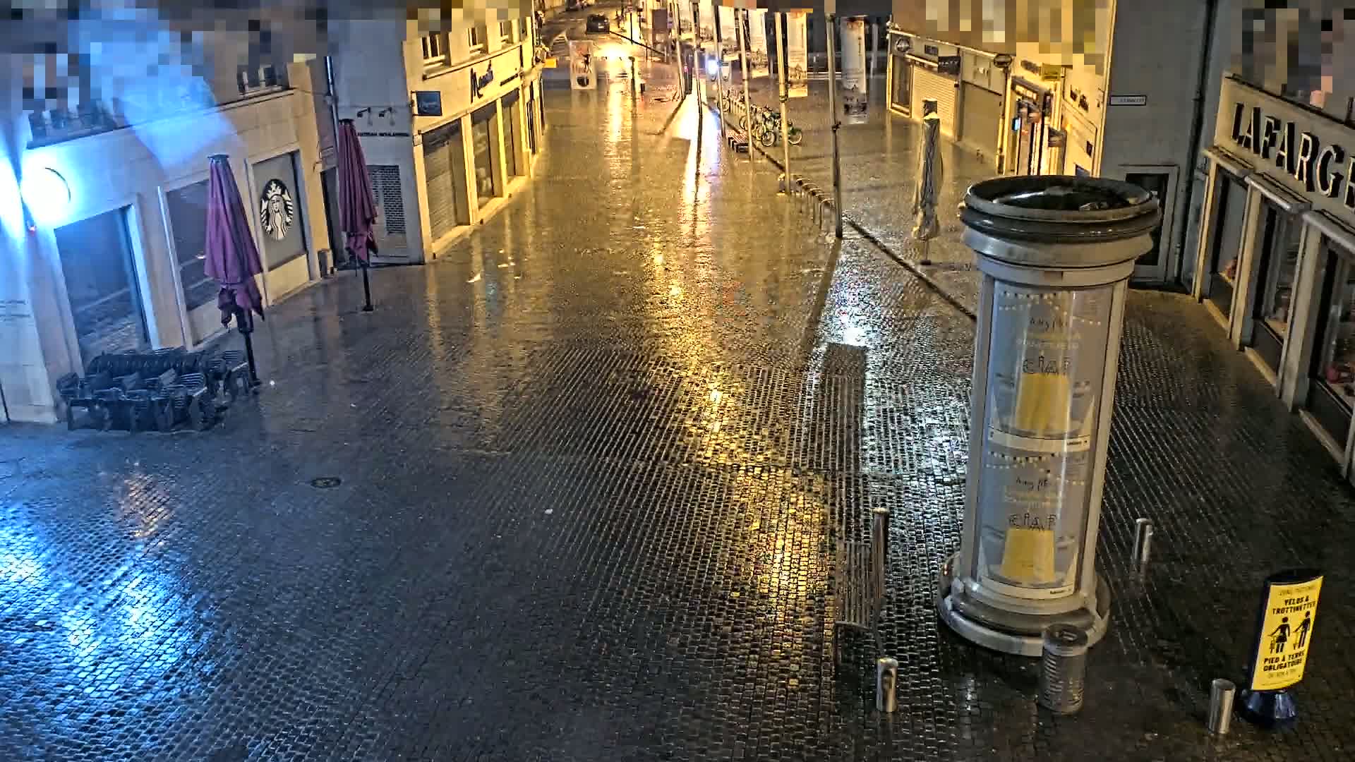 A wet, cobblestone street at night, illuminated by streetlights, with closed shops and outdoor seating areas visible.