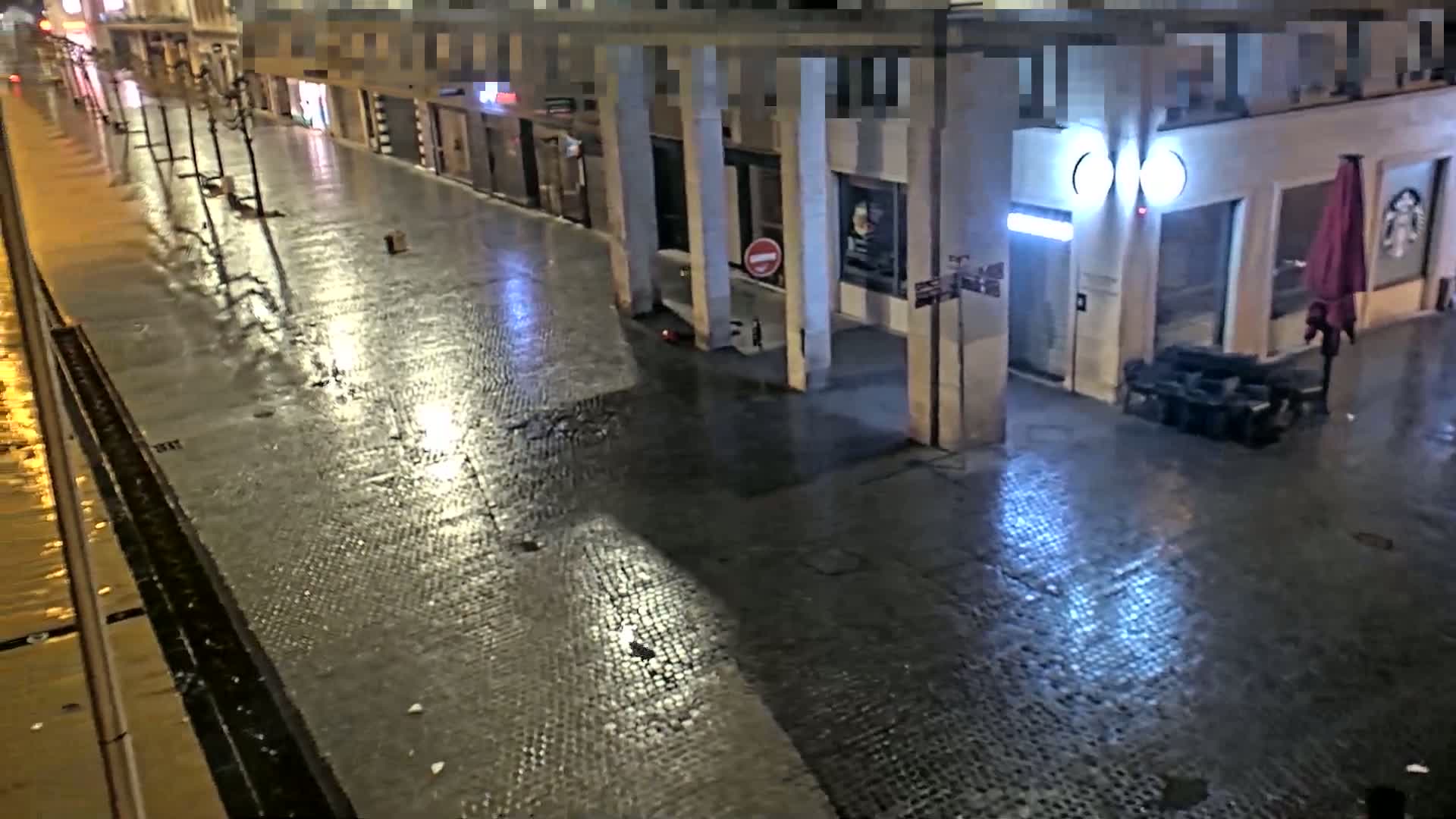 A rain-slicked cobblestone street at night, lined with buildings and lampposts, reflects the ambient light.