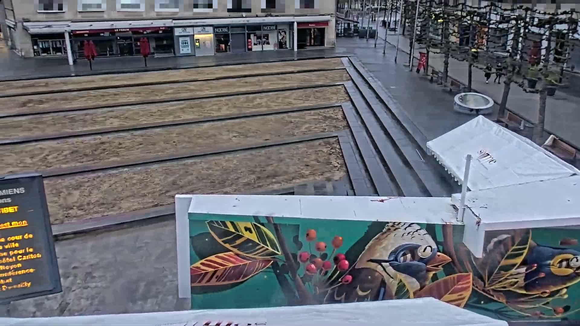 A partially constructed urban plaza with sand-filled tram tracks, steps, and a mural depicting birds and foliage is visible on an overcast day.