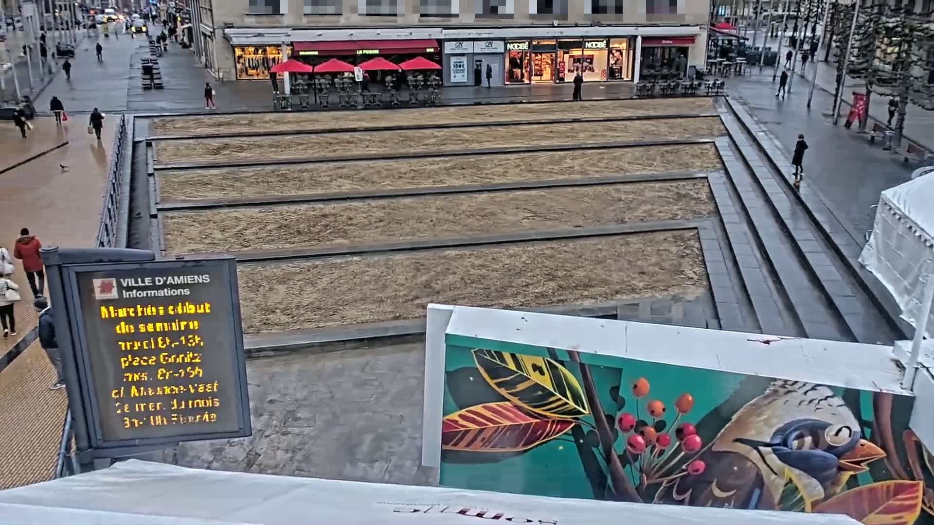 A mostly empty paved square with long, narrow, sandy sections, viewed from above on a cloudy day.