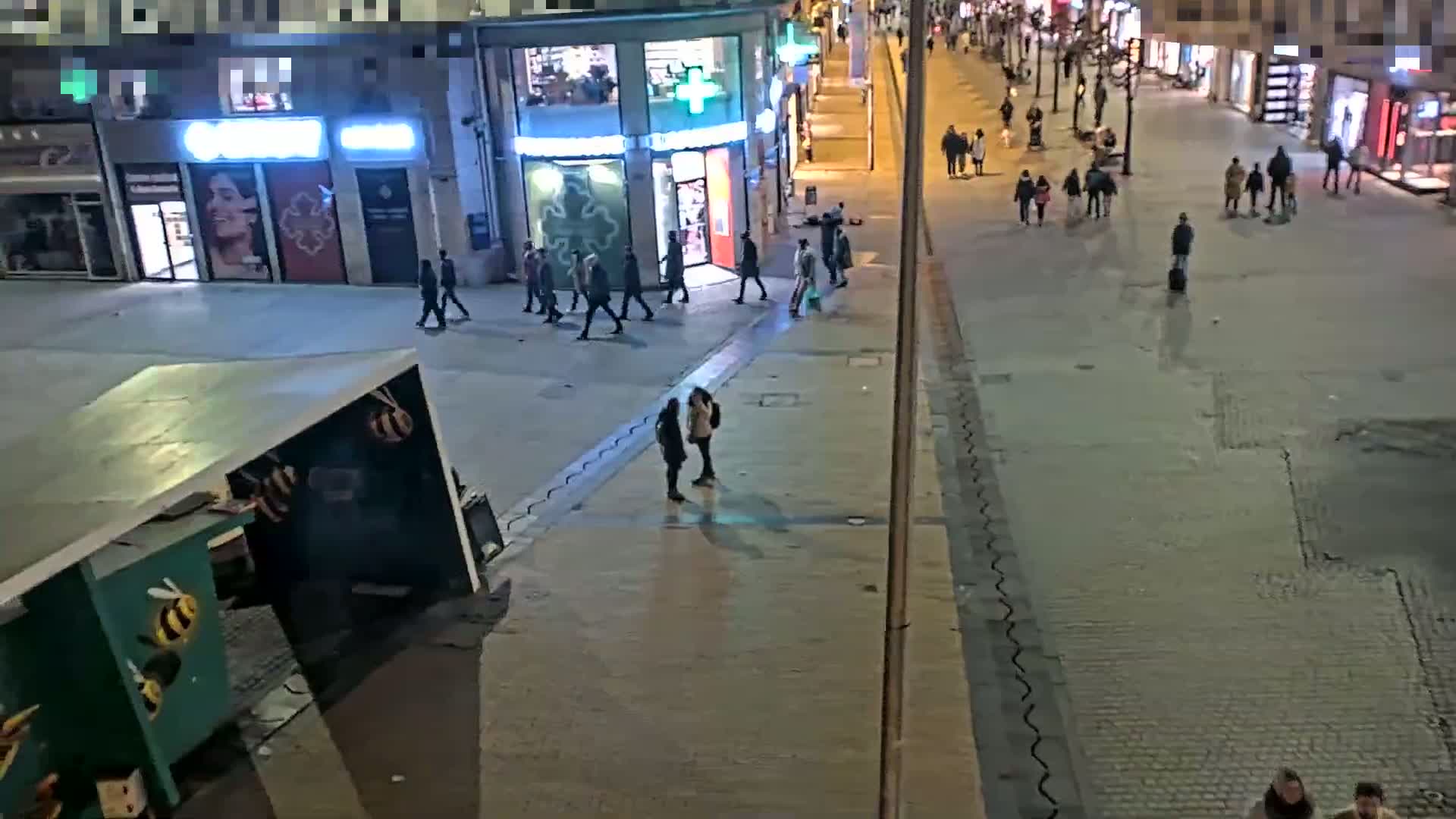 A nighttime, outdoor street scene shows sparsely populated sidewalks and a street with pedestrians walking past shops and buildings.