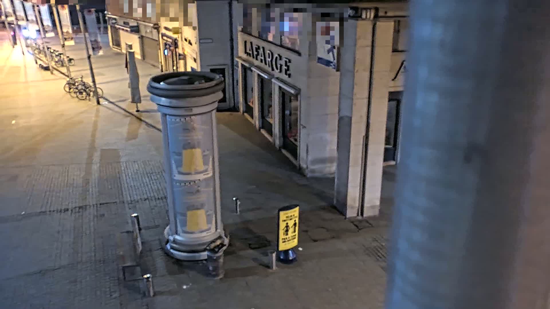 A nighttime street scene shows a cylindrical advertising column, a few parked bicycles, and a storefront with a yellow social distancing sign.