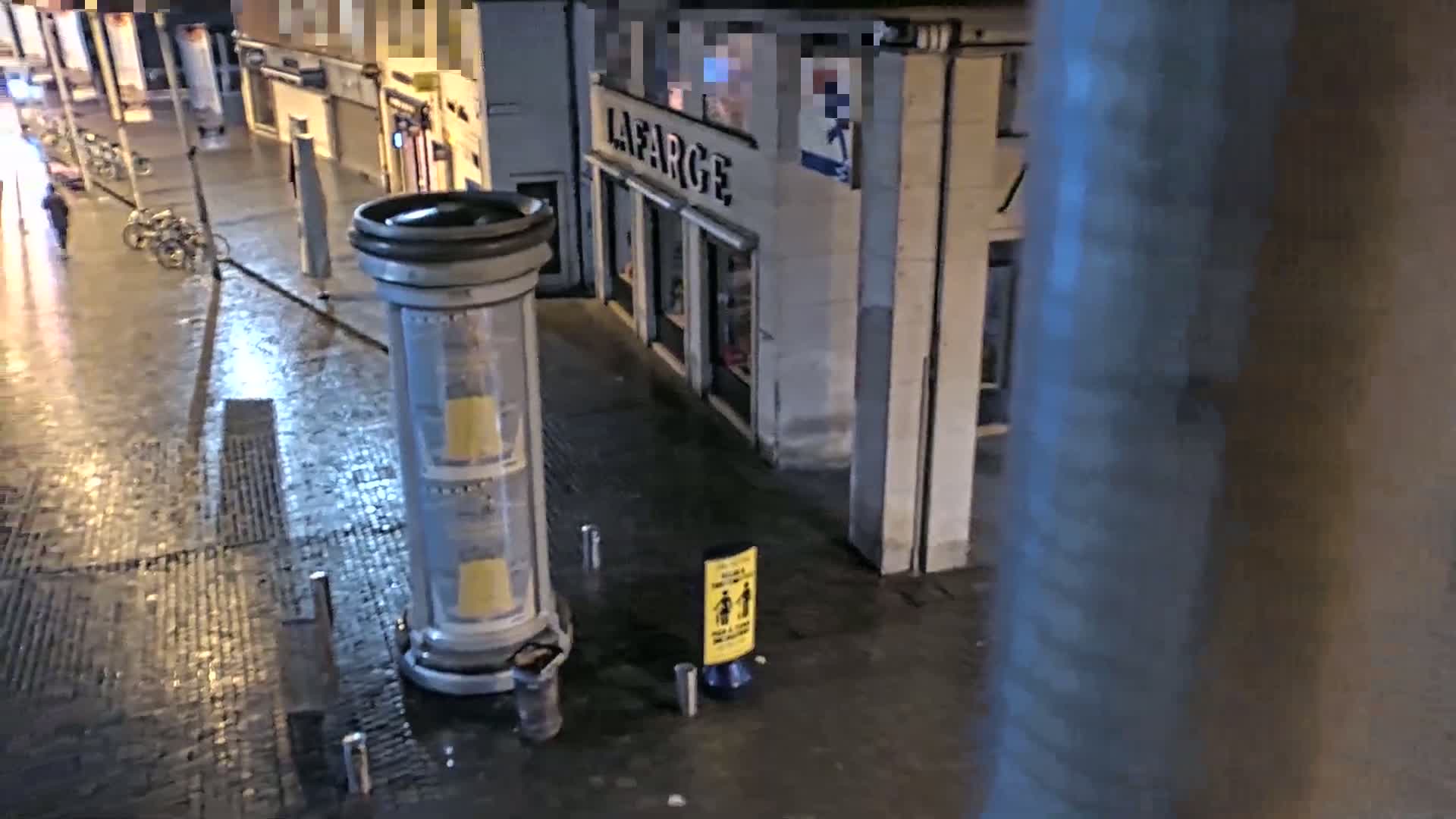 A wet, nighttime street scene shows a cylindrical advertisement pillar near parked bicycles and a building with a store named Lafarge.