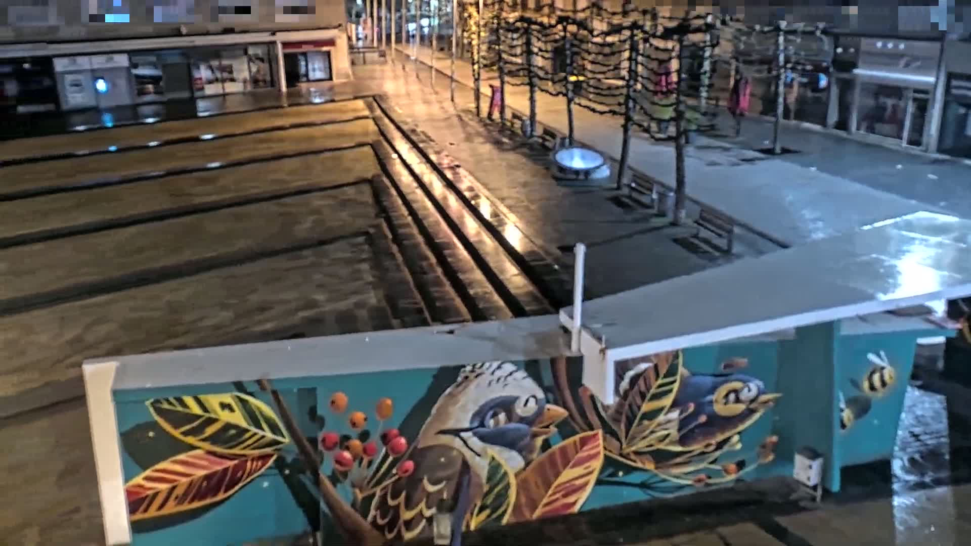 A wet, nighttime city plaza features a mural of birds and leaves on a structure overlooking a multi-tiered water feature.