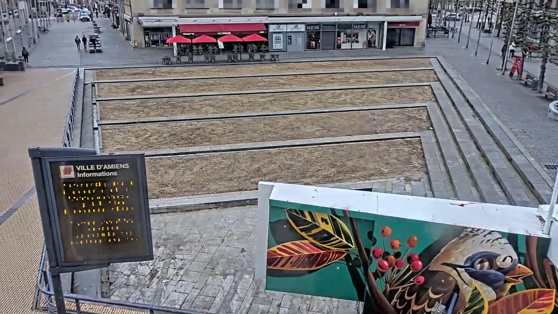 A paved plaza with three long, rectangular, bare dirt sections, overlooked by buildings and a street with sparse pedestrian traffic, under an overcast sky.