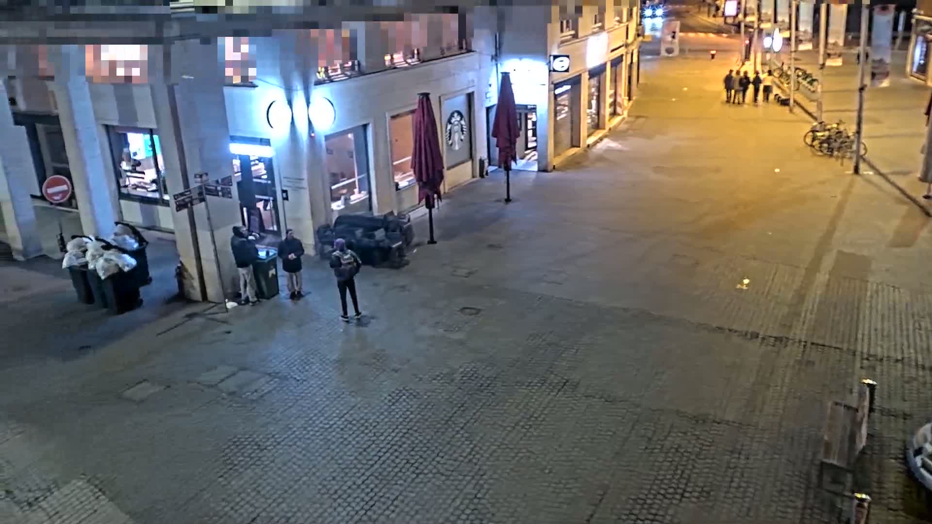 At night, several people stand near a Starbucks and some garbage bins on a cobblestone street, under outdoor lighting.