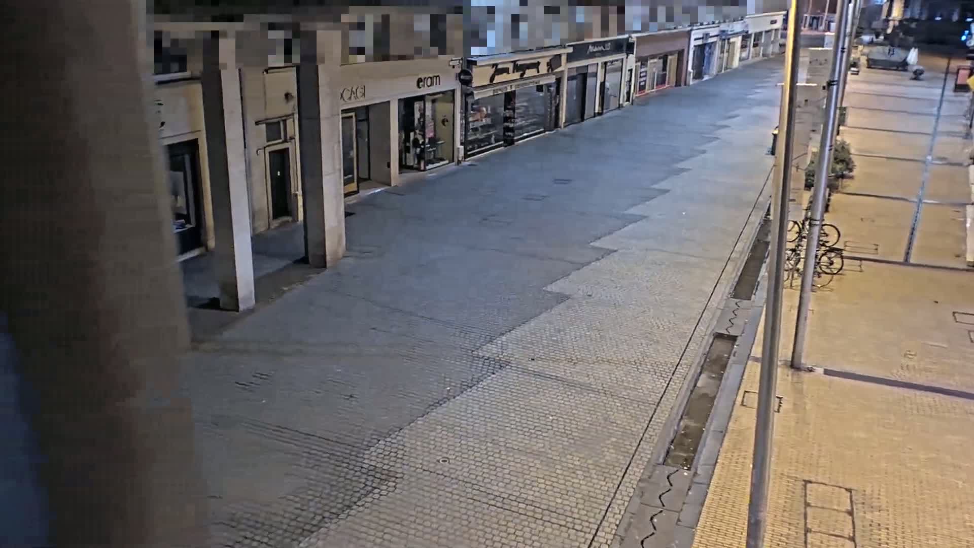 A mostly empty, paved street at night with shops lining one side, illuminated by streetlights, and a few bicycles parked nearby.