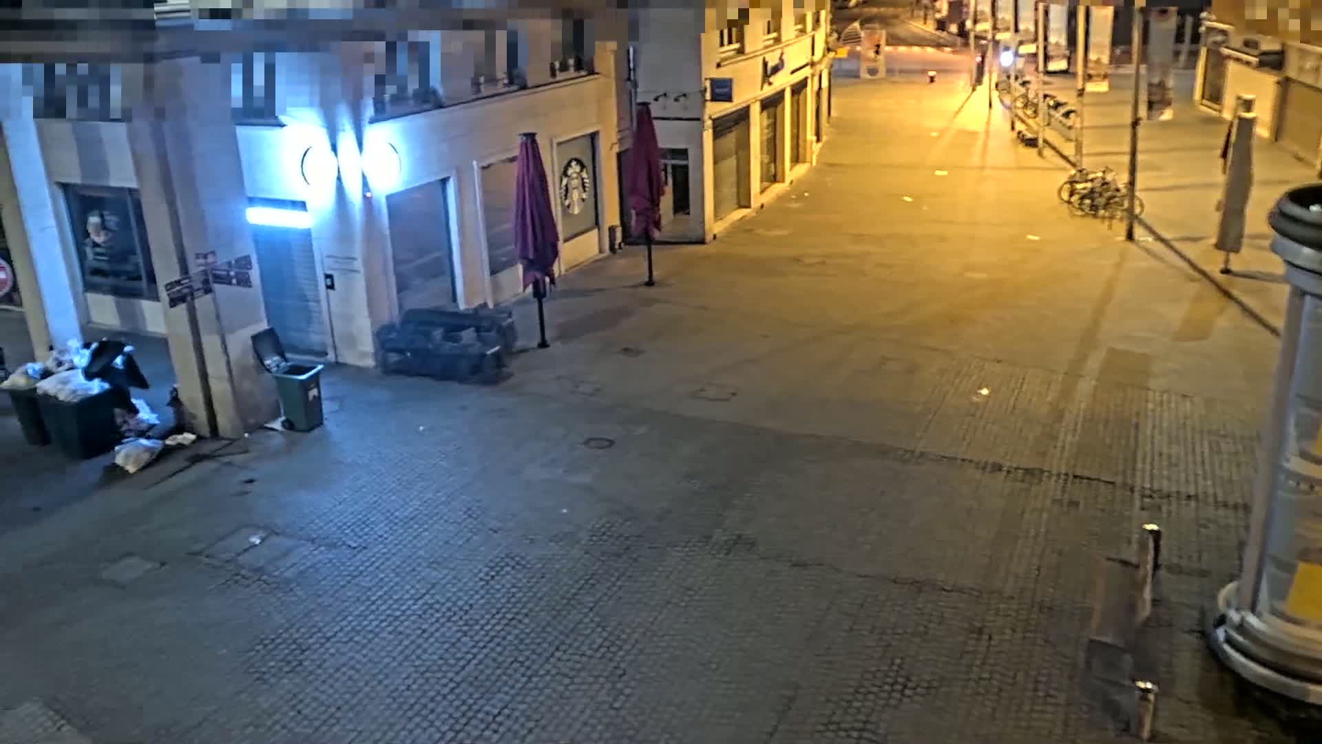 An empty city street at night, illuminated by streetlights, shows overflowing trash cans near closed shops with umbrellas folded outside.