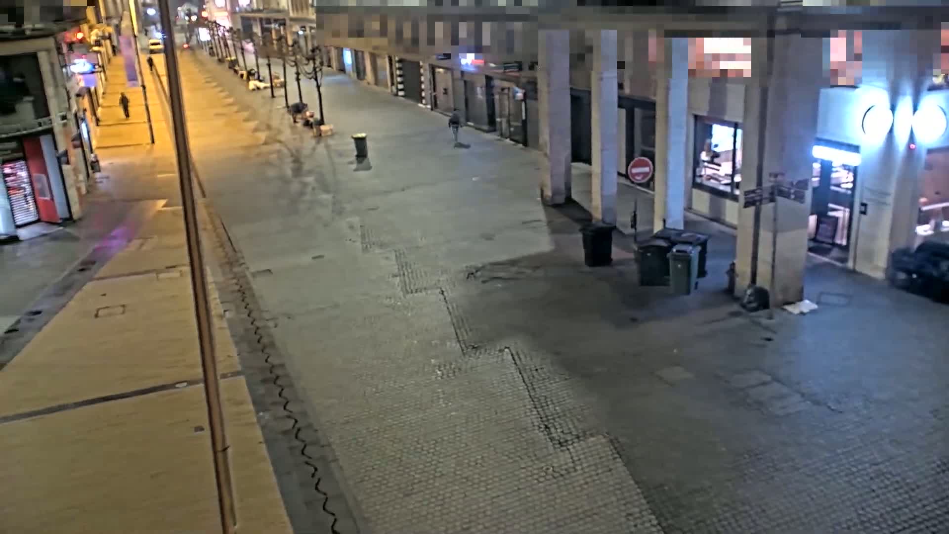 A mostly empty city street at night is lit by streetlights, with a few pedestrians visible and some trash cans near buildings.