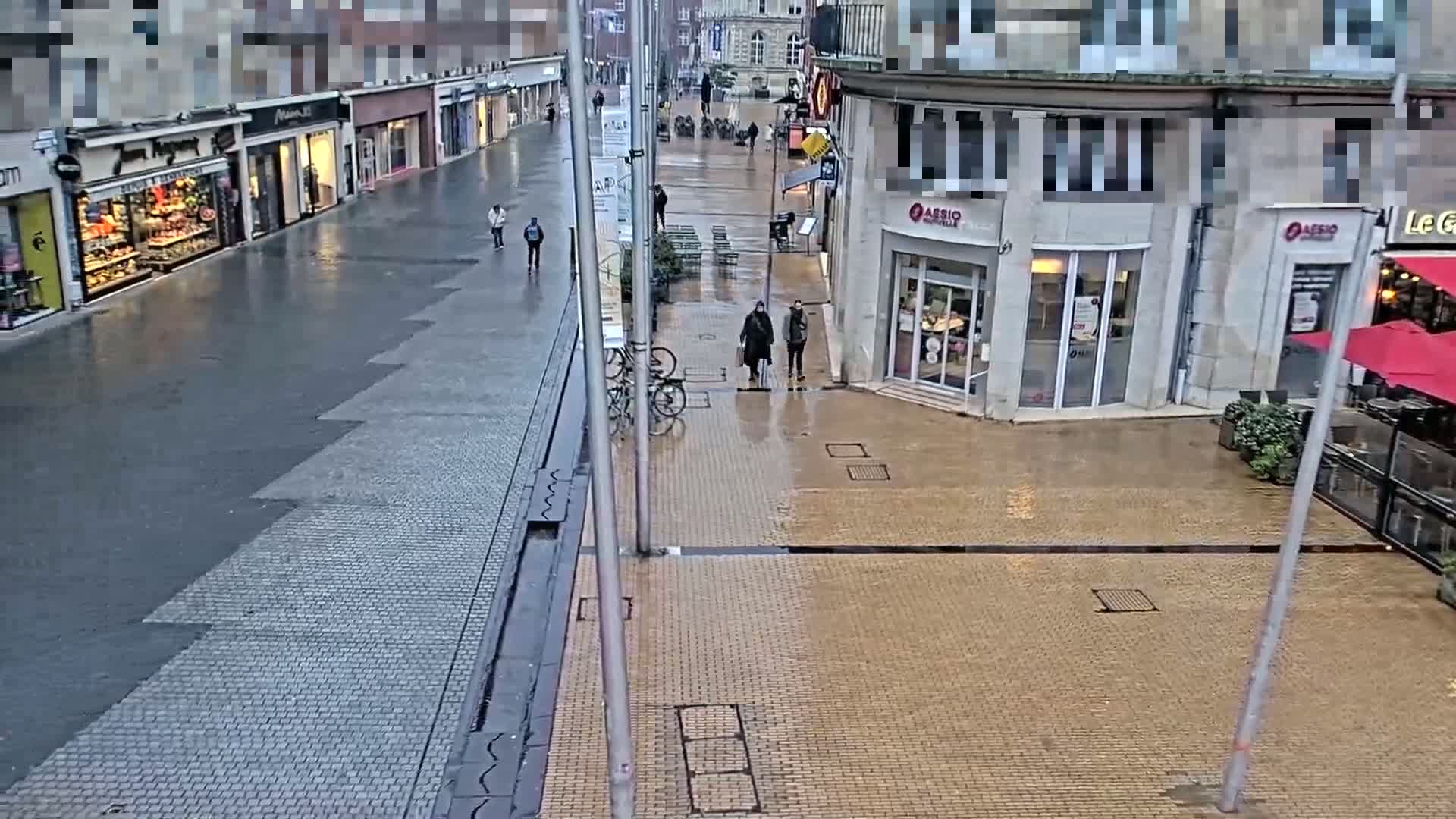 A wet, paved city street scene shows a few pedestrians and some parked bicycles near shops and restaurants on a rainy day.