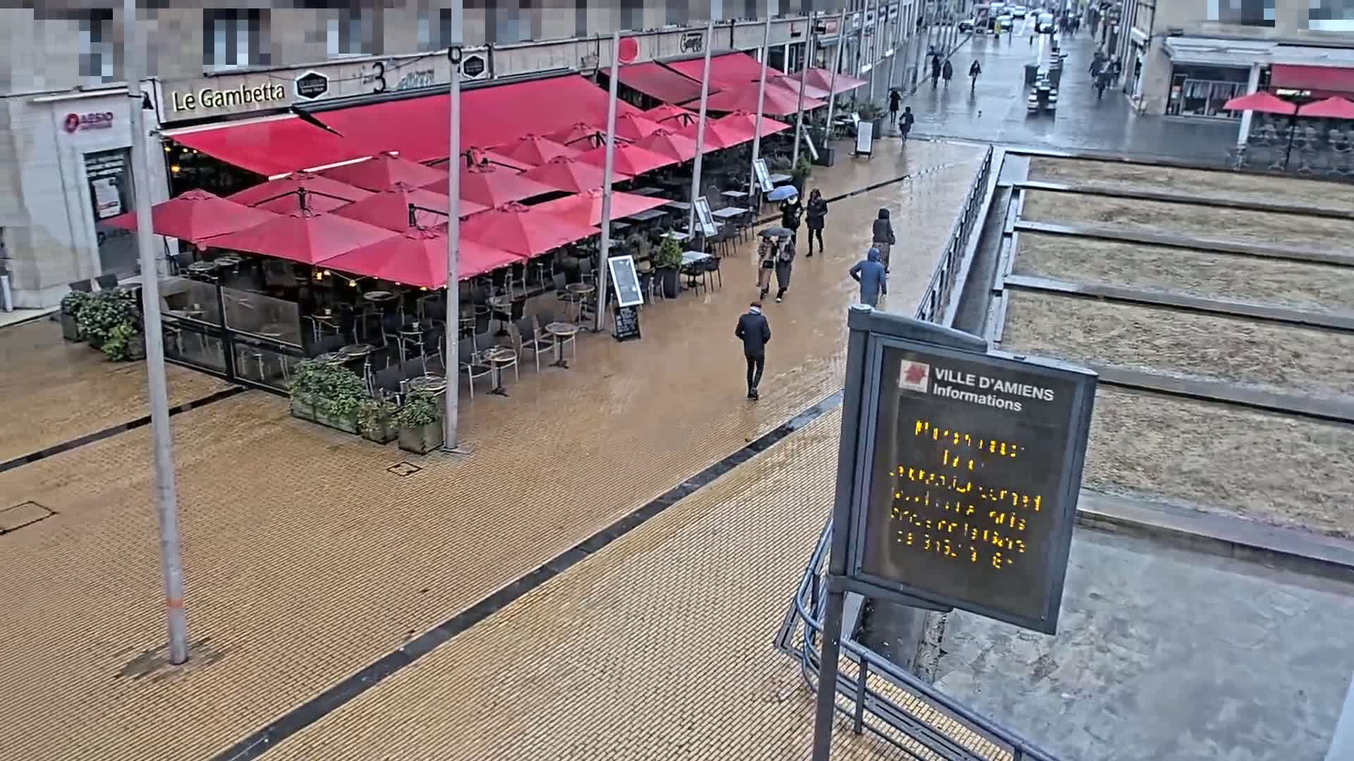 A wet, paved city street features a restaurant with red umbrellas and several people walking under an overcast sky.