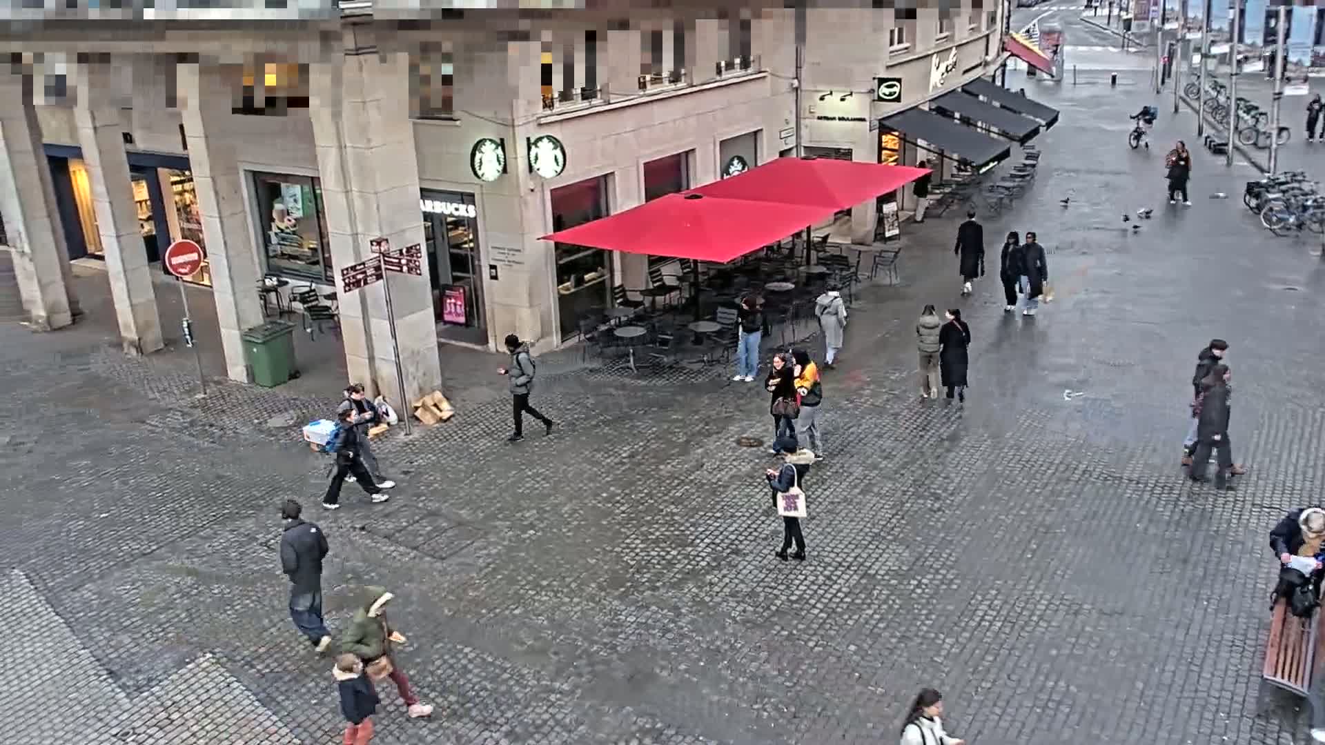 A wet cobblestone plaza features a Starbucks with a red awning and several people walking around.
