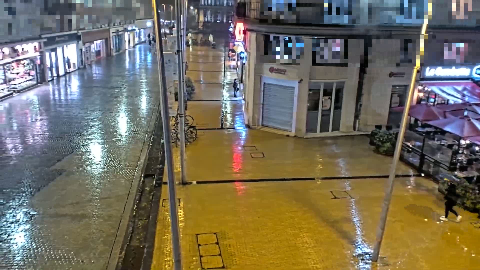 A rain-slicked city street at night, with shops and a restaurant visible, shows reflections of lights on the wet pavement.