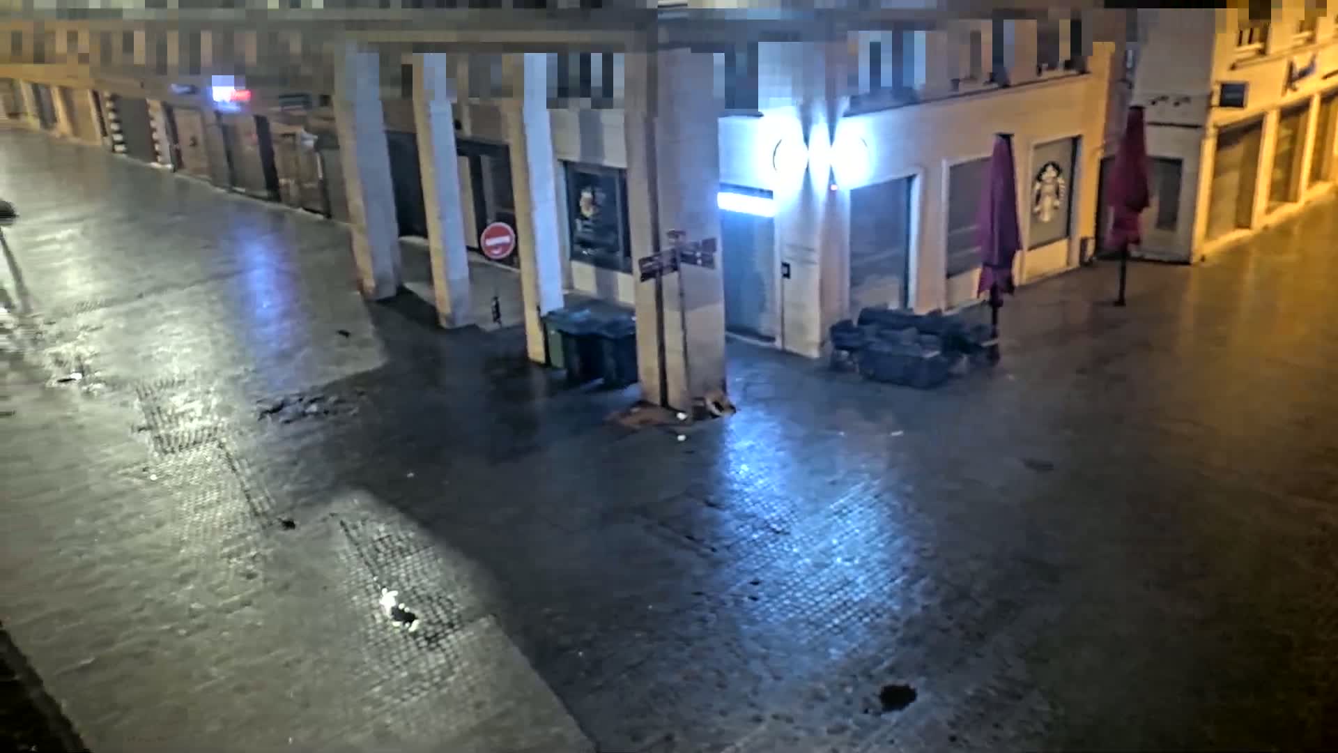 A wet, nighttime street scene shows a mostly empty alleyway between buildings with closed storefronts and a few closed umbrellas.