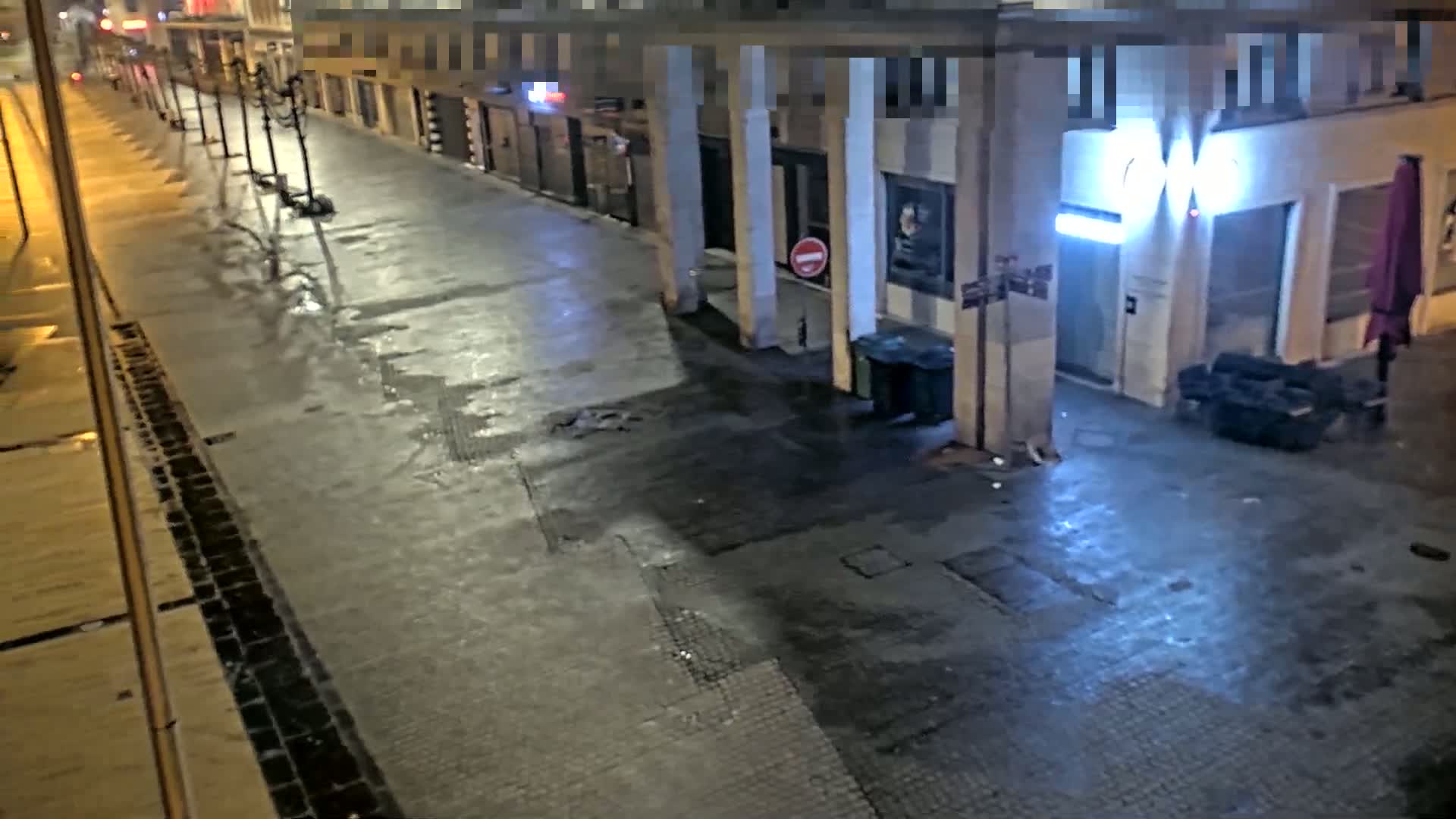 A wet, empty street at night is lined with buildings, some with illuminated signs.