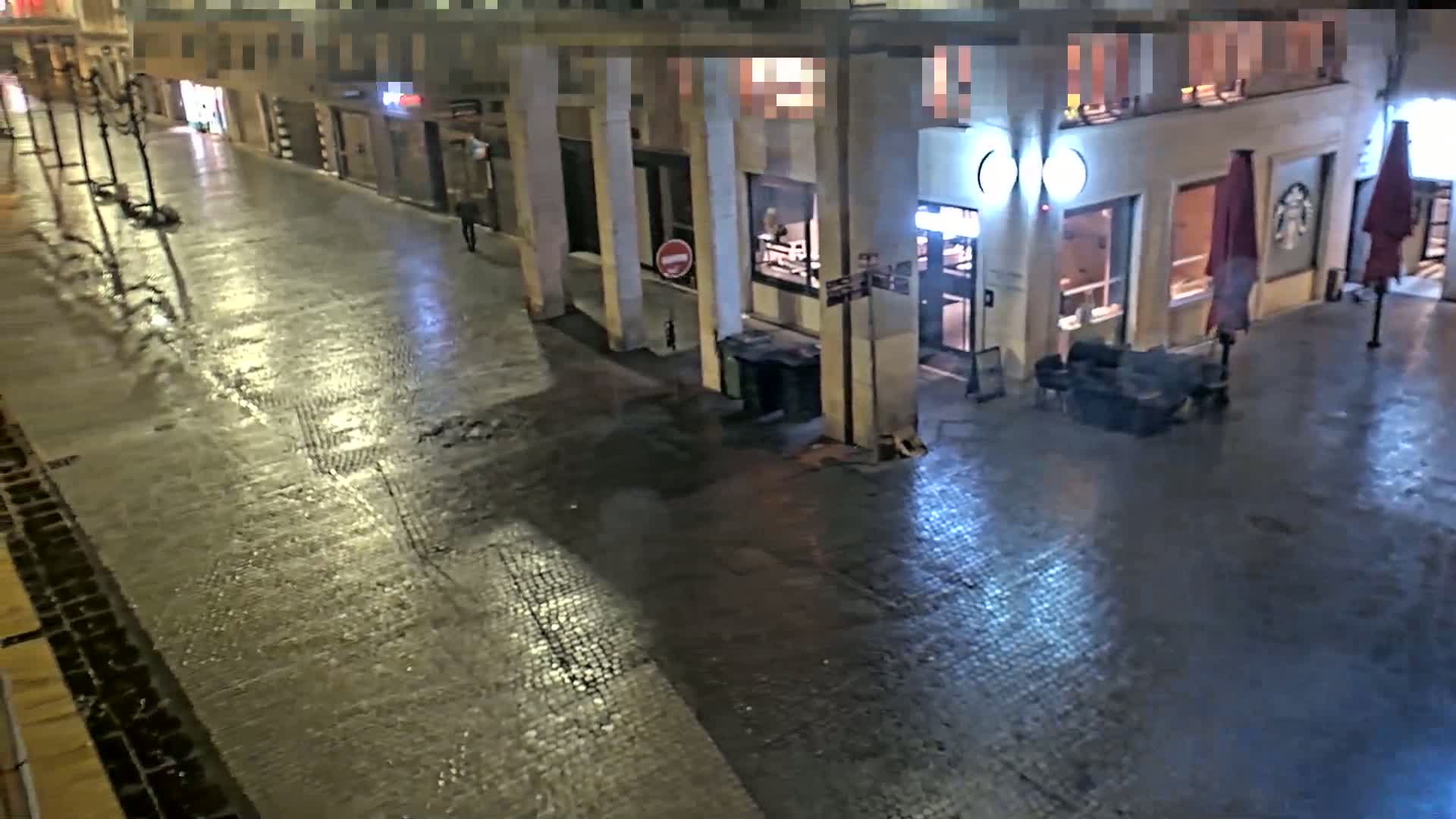 A wet, empty city street at night, illuminated by streetlights, with a building featuring outdoor seating and closed storefront visible.
