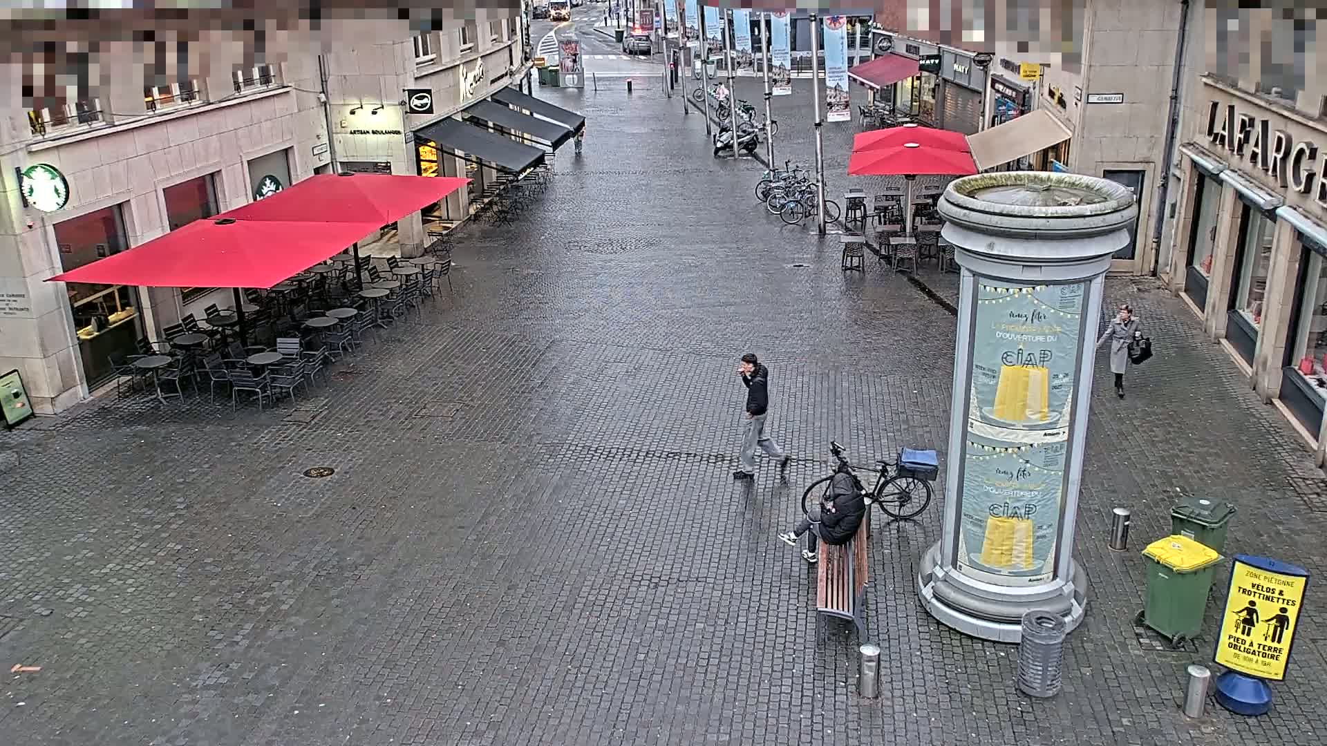 A wet cobblestone street scene shows several people, some sitting on a bench near a bicycle, with red umbrellas shading outdoor cafe seating.