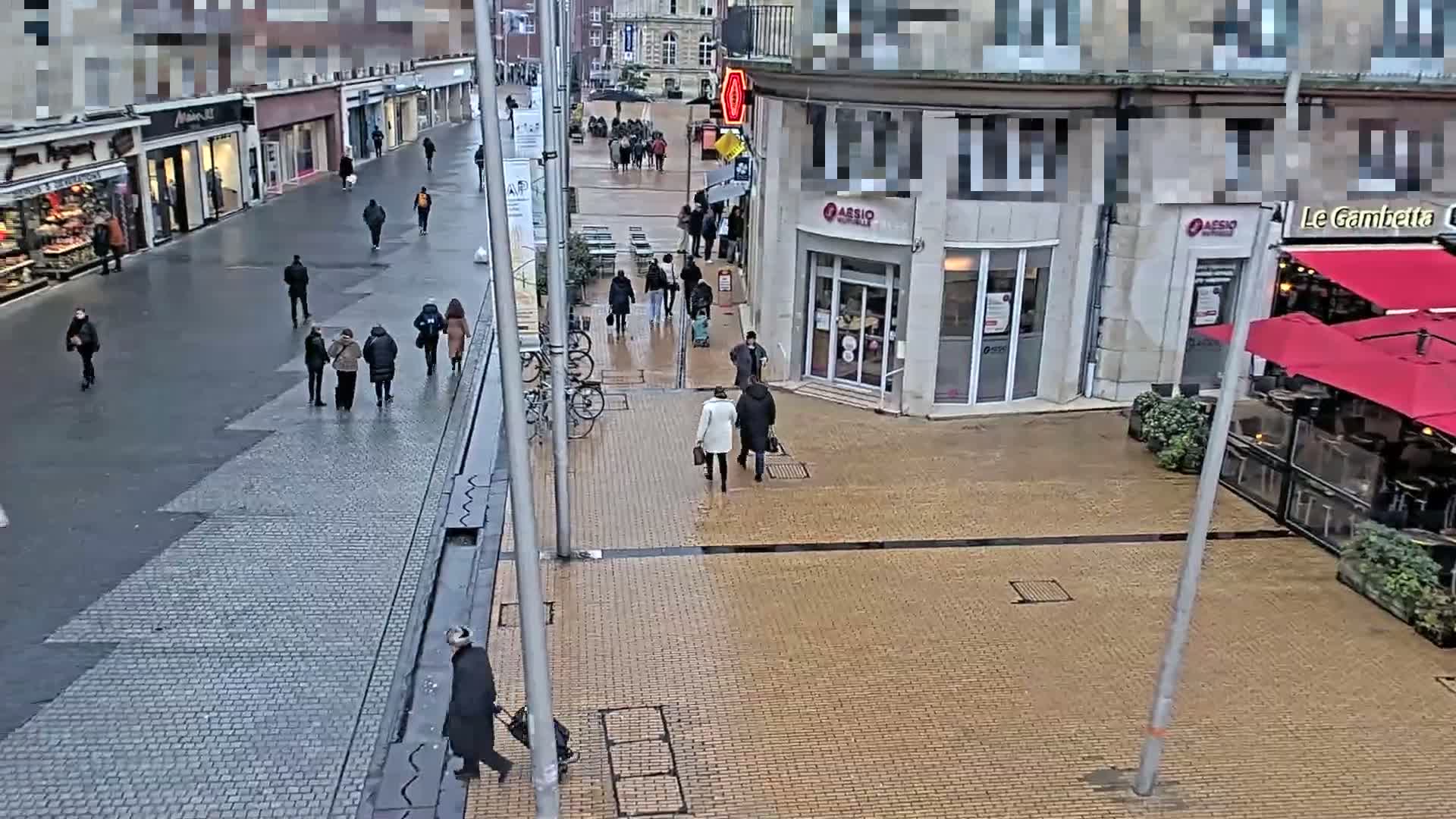 A wet, paved pedestrian area with several people walking past shops and a restaurant with red umbrellas.
