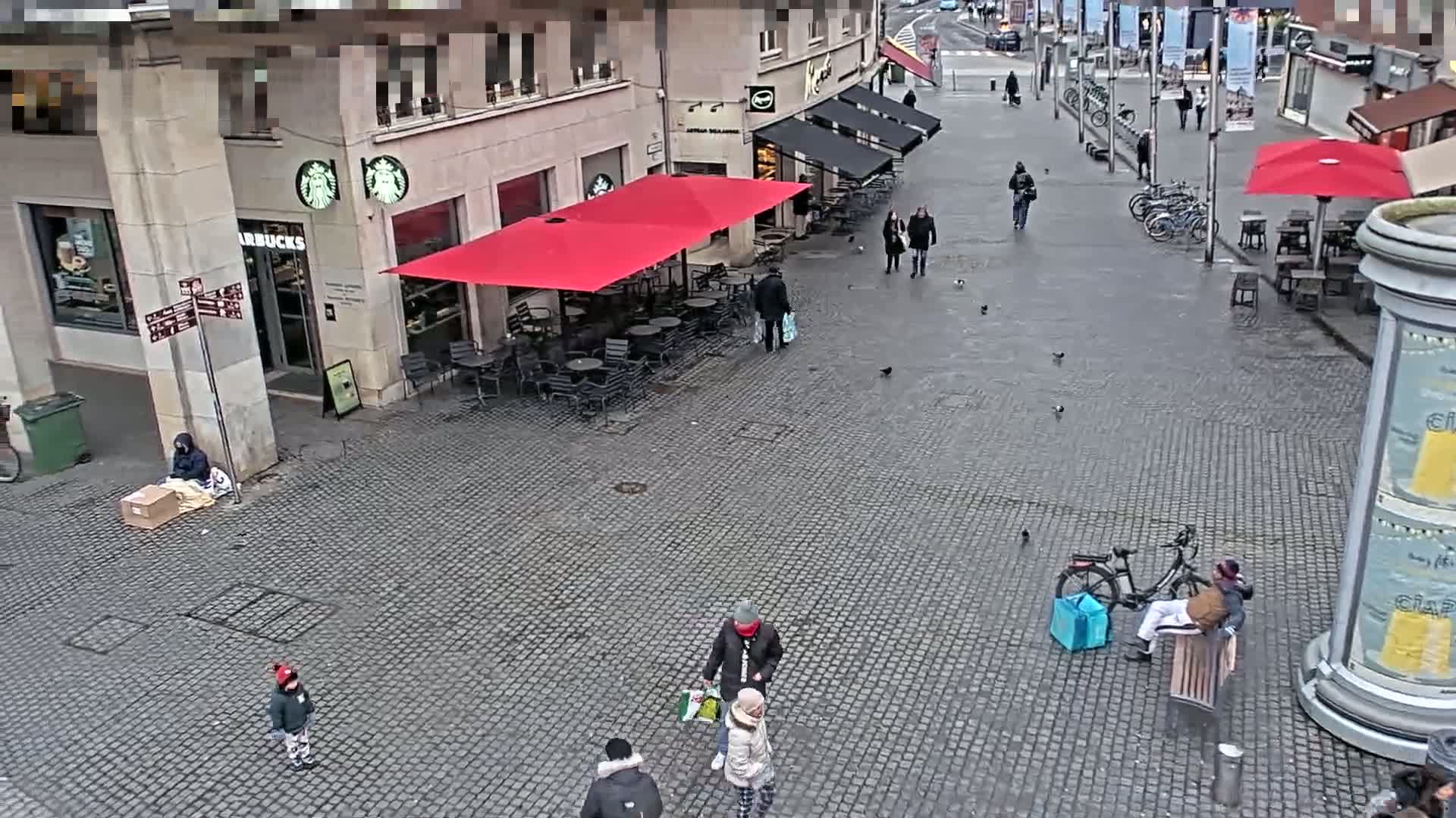 A cobblestone plaza with several people walking and sitting, some outdoor seating areas with red and black umbrellas, and a person sitting on the ground next to a box on a cloudy day.