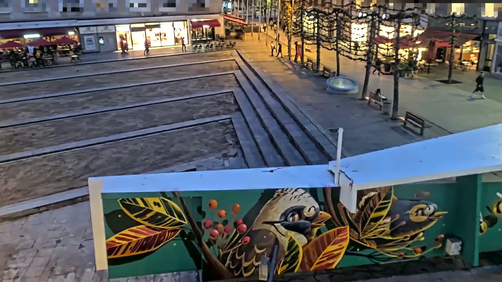 A nighttime view of a city square with a mural of birds and leaves on a wall, showing a paved area with some people walking around.