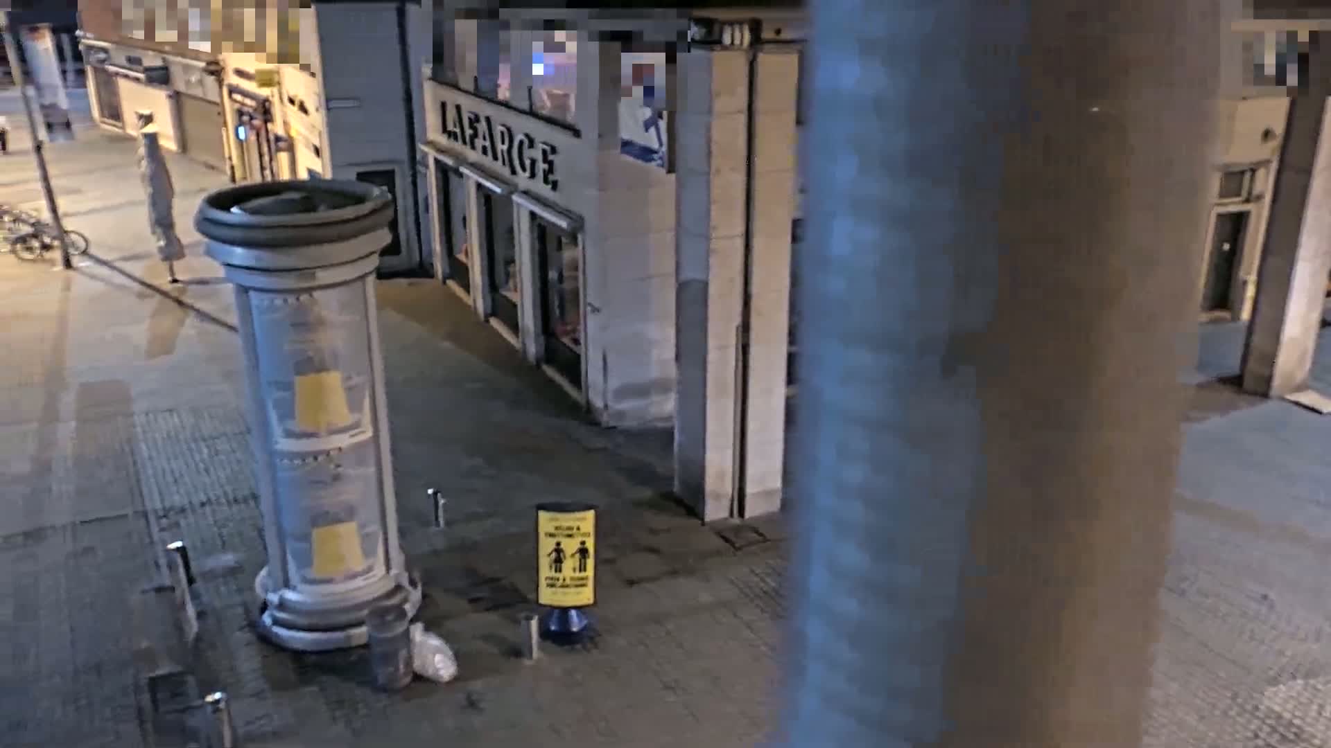 A nighttime street scene shows a mostly empty sidewalk, a large cylindrical advertising column, a store, and a social distancing sign.
