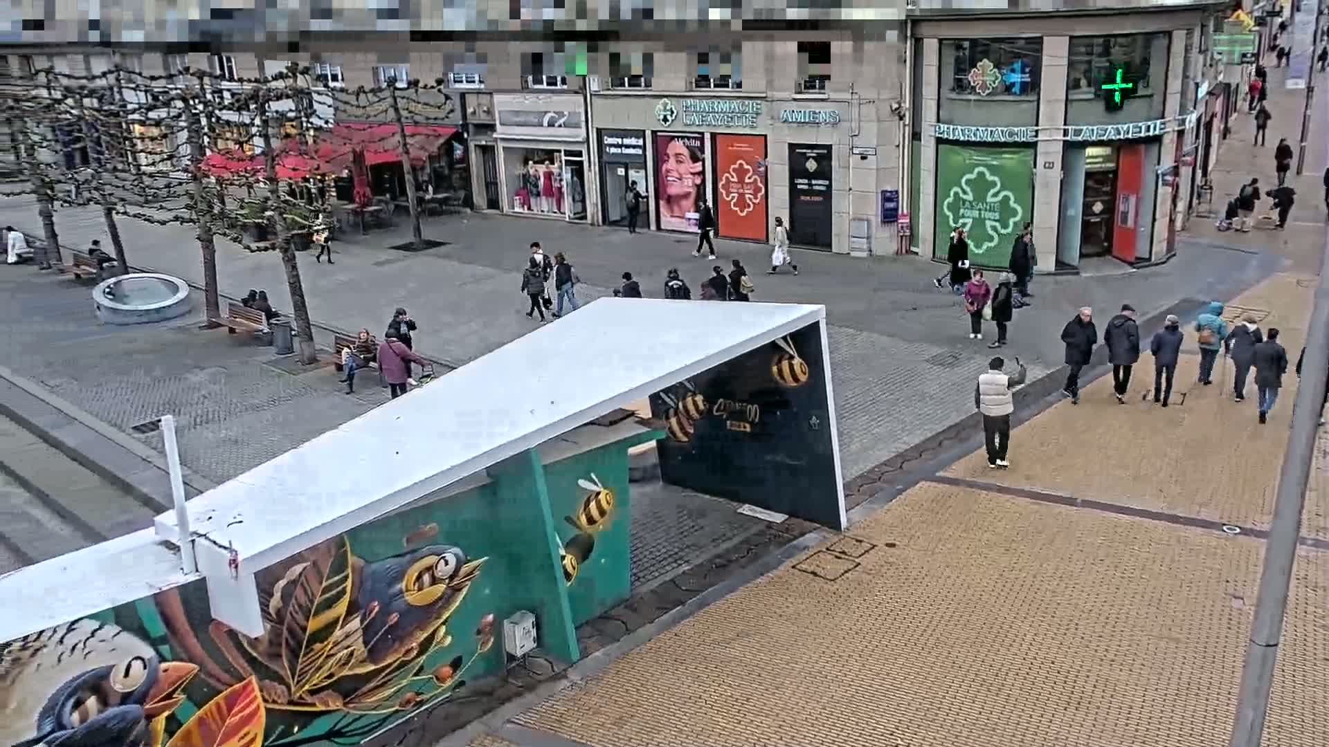 A city street scene on an overcast day shows pedestrians walking past shops and a structure with a mural depicting birds and bees.