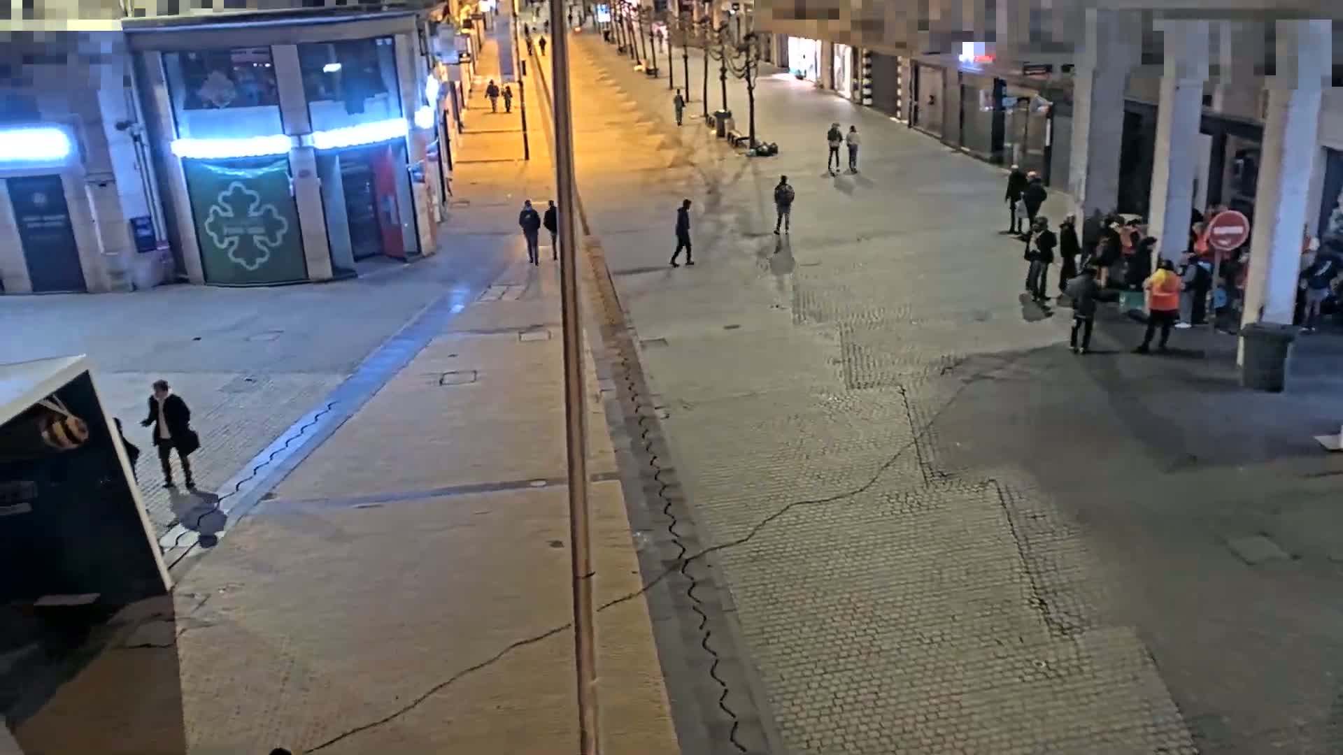 A mostly empty city street at night, illuminated by streetlights, shows several small groups of people standing and walking.