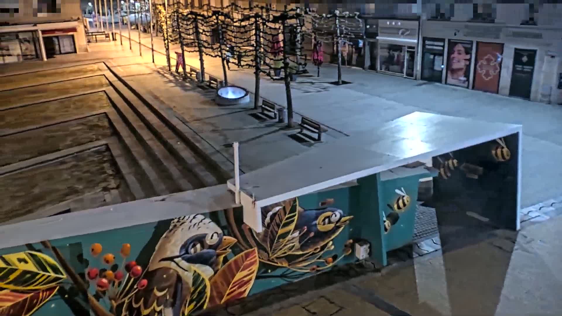 A nighttime view of a city plaza with a mural of birds and bees on a wall, under a partially covered walkway, near a water feature and shops.