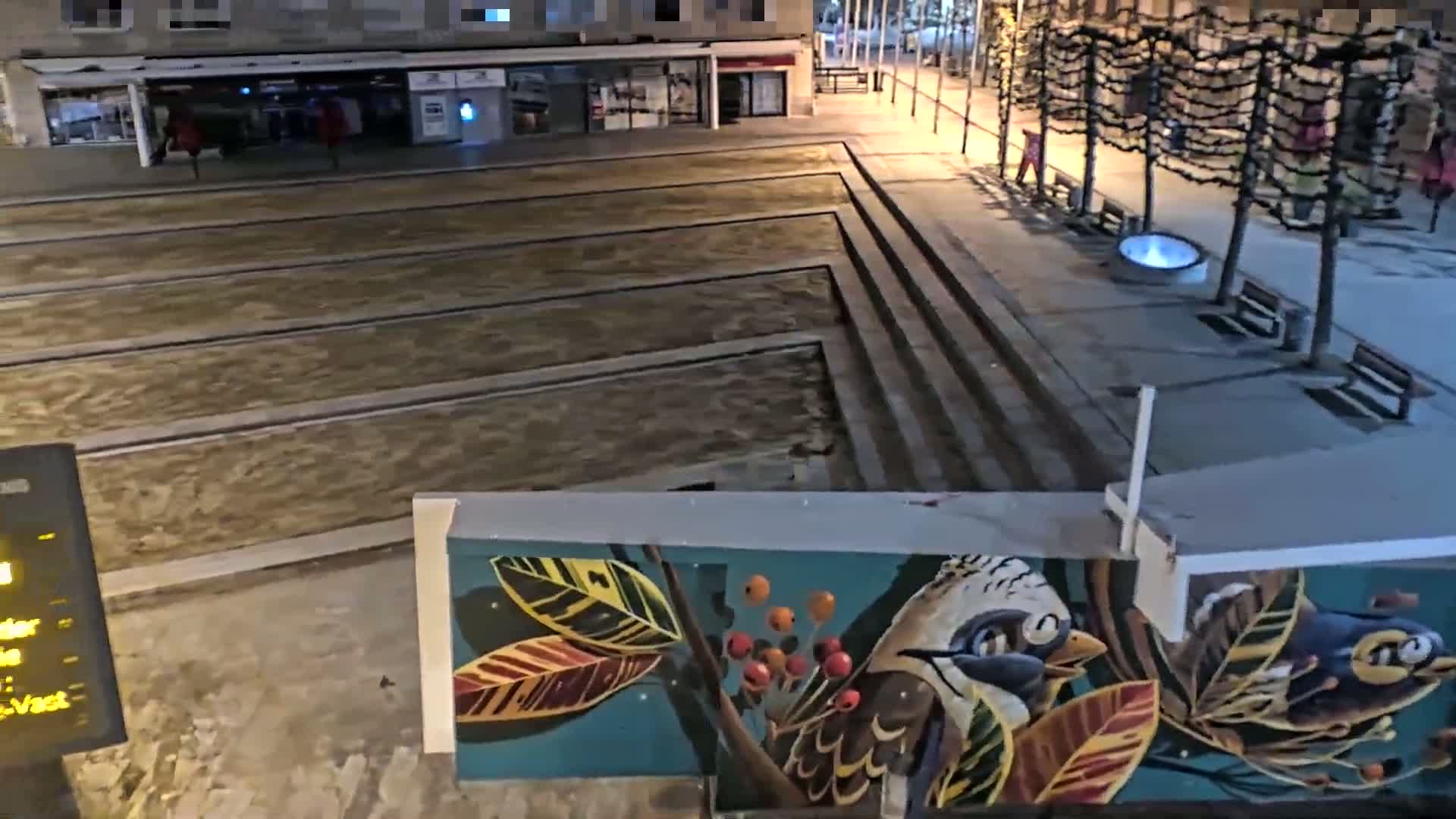 A nighttime view of a city plaza with a mural depicting birds and foliage, featuring a sunken, rectangular water feature.