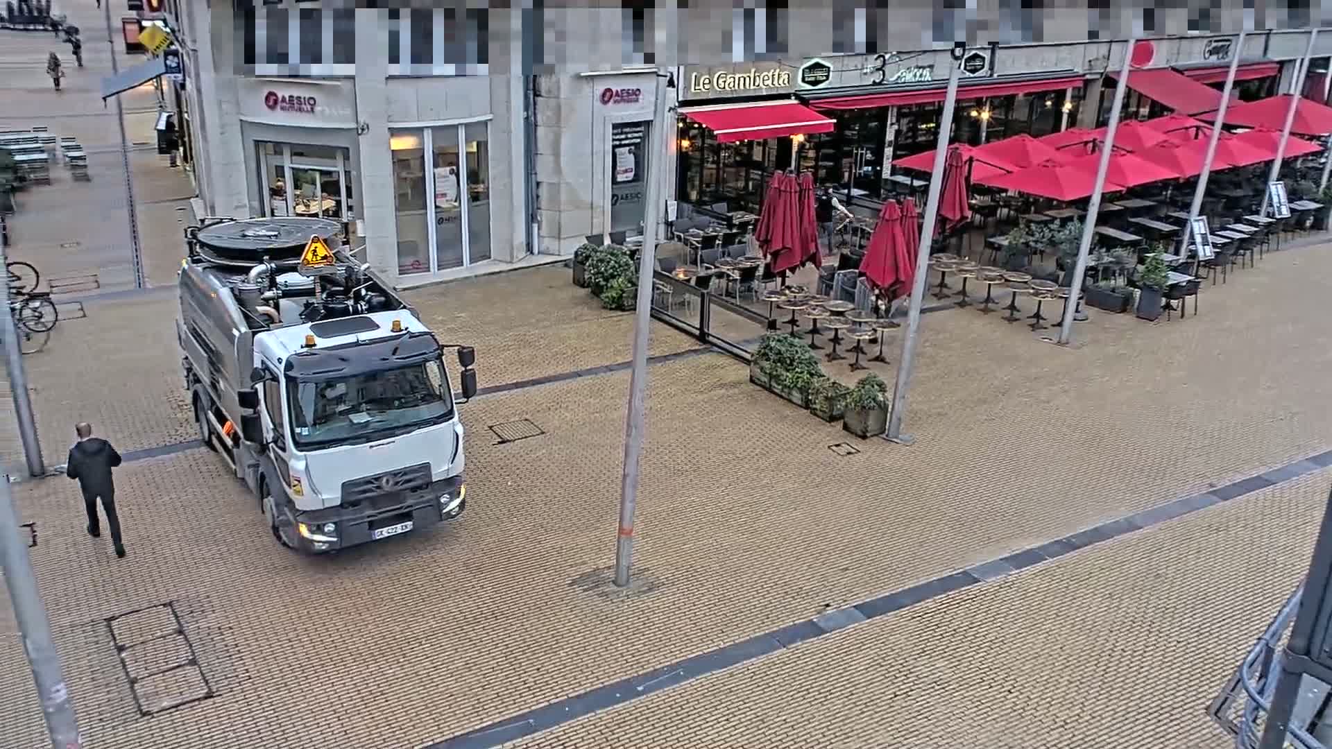 A white vacuum truck is parked on a brick-paved street next to an outdoor cafe with red umbrellas under an overcast sky.