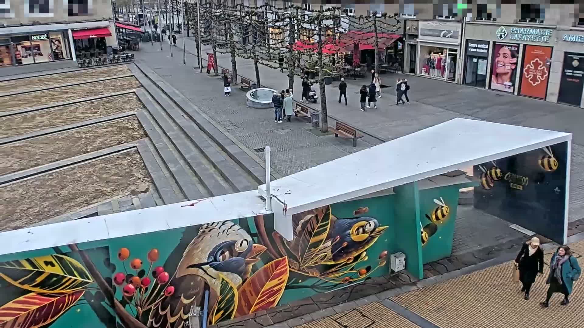 A city square with a mural of birds and bees on a structure, partially filled with dirt, is viewed from above on an overcast day with several people walking around.
