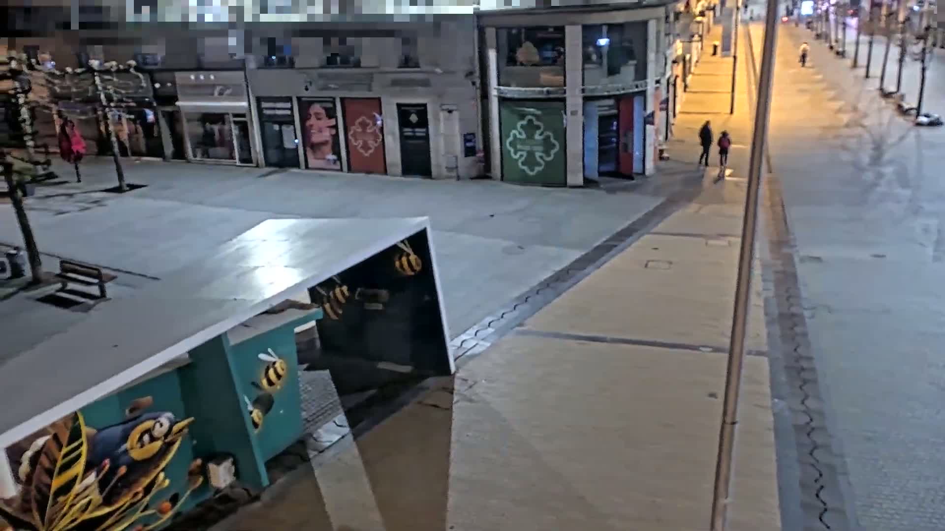 A nighttime, city street scene shows a mostly empty plaza with shops and a mural of bees on a structure.