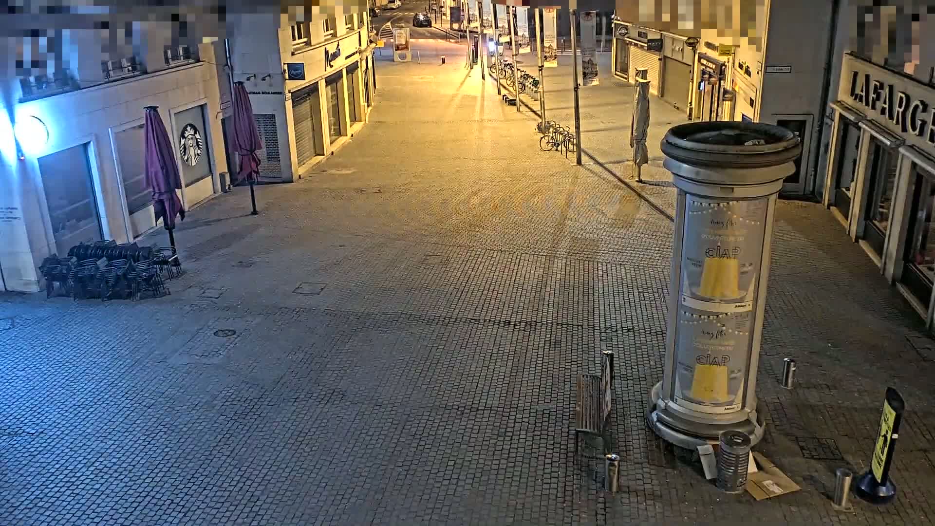 A nearly deserted cobblestone street at night is lit by streetlights, with a few parked bicycles and a bench visible.