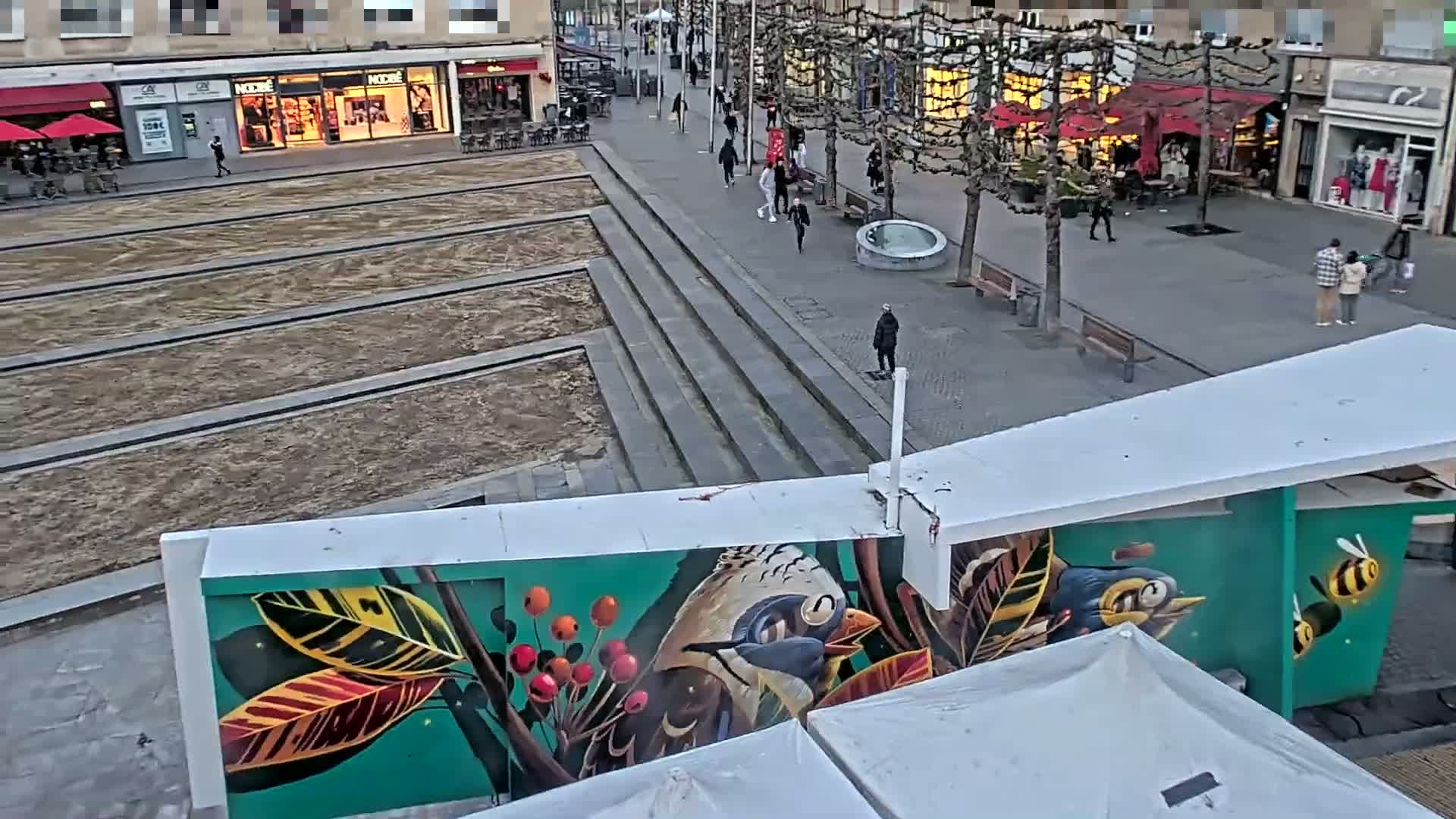 An overhead view shows a city square under a cloudy sky, with pedestrians walking past shops and a large mural depicting birds and leaves on a building.