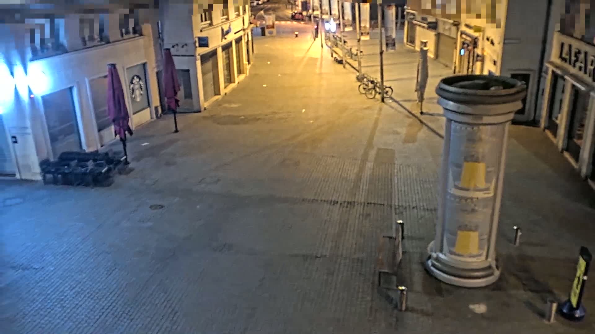 A mostly empty city street at night is illuminated by streetlights, with a bicycle parked near a cylindrical advertising column and closed storefronts along the sides.