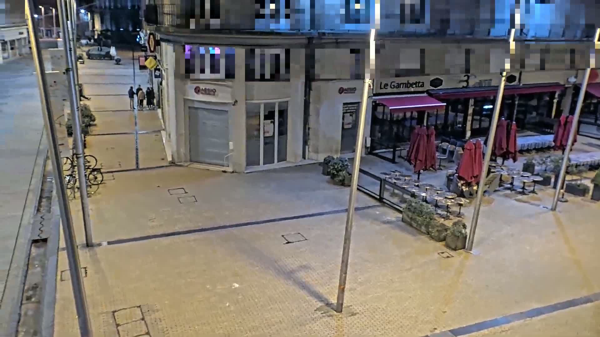 At night, a mostly empty plaza with a restaurant featuring closed red umbrellas and several bicycles parked nearby.