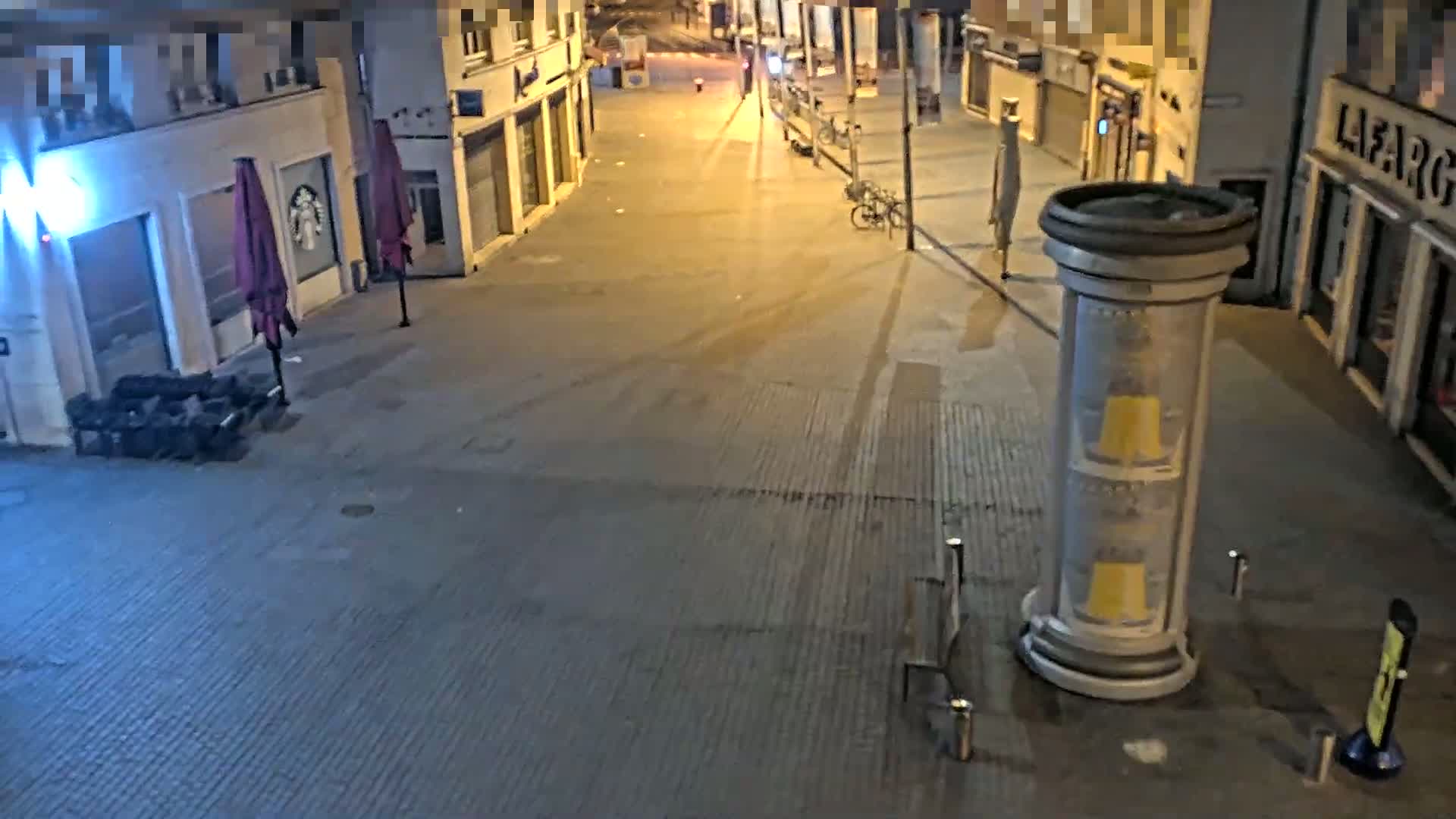 An empty street at night, lit by streetlights, shows closed shops and a few pieces of outdoor furniture.