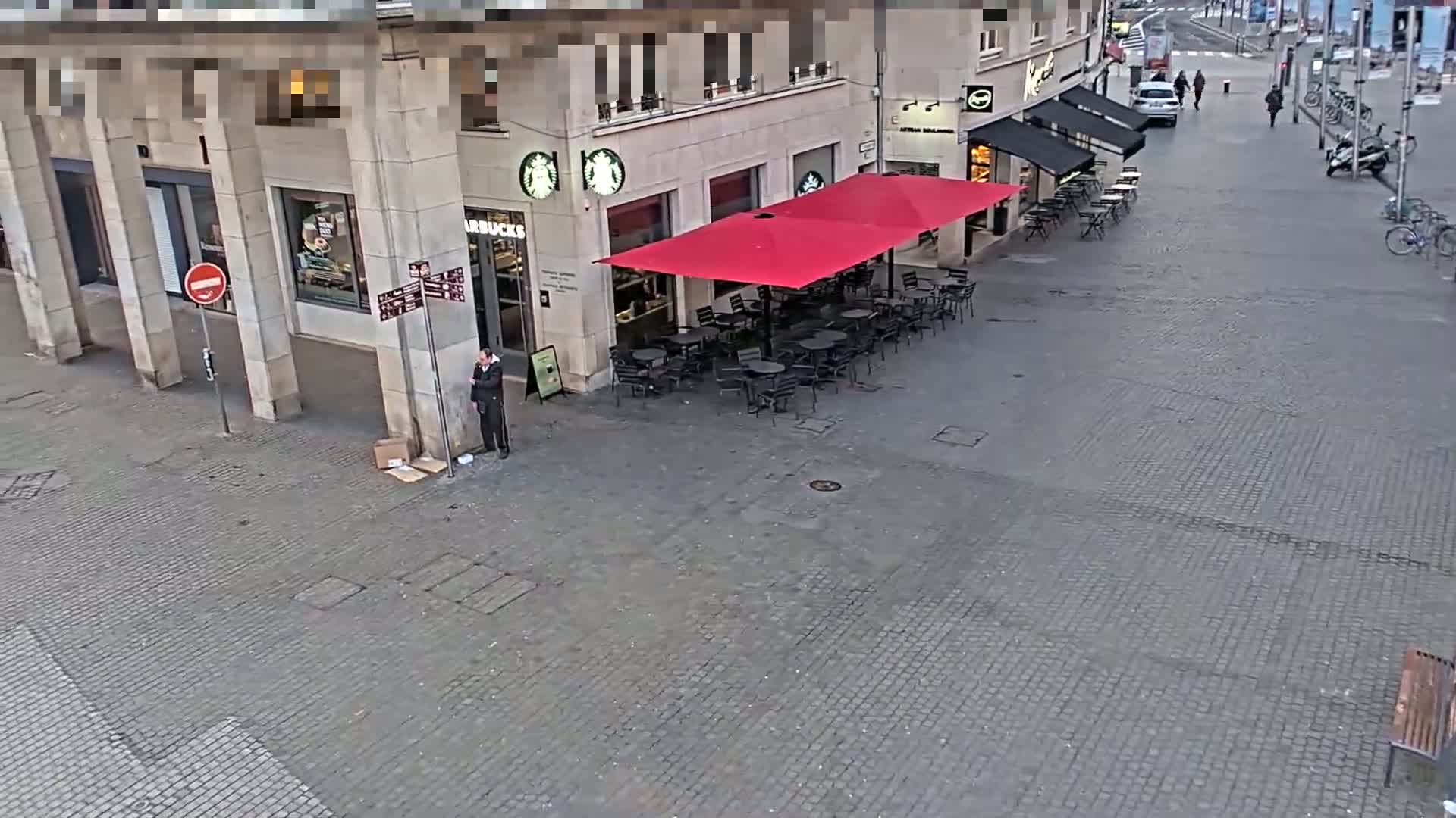 A person stands near a Starbucks with an outdoor seating area under a red umbrella on a paved city square.