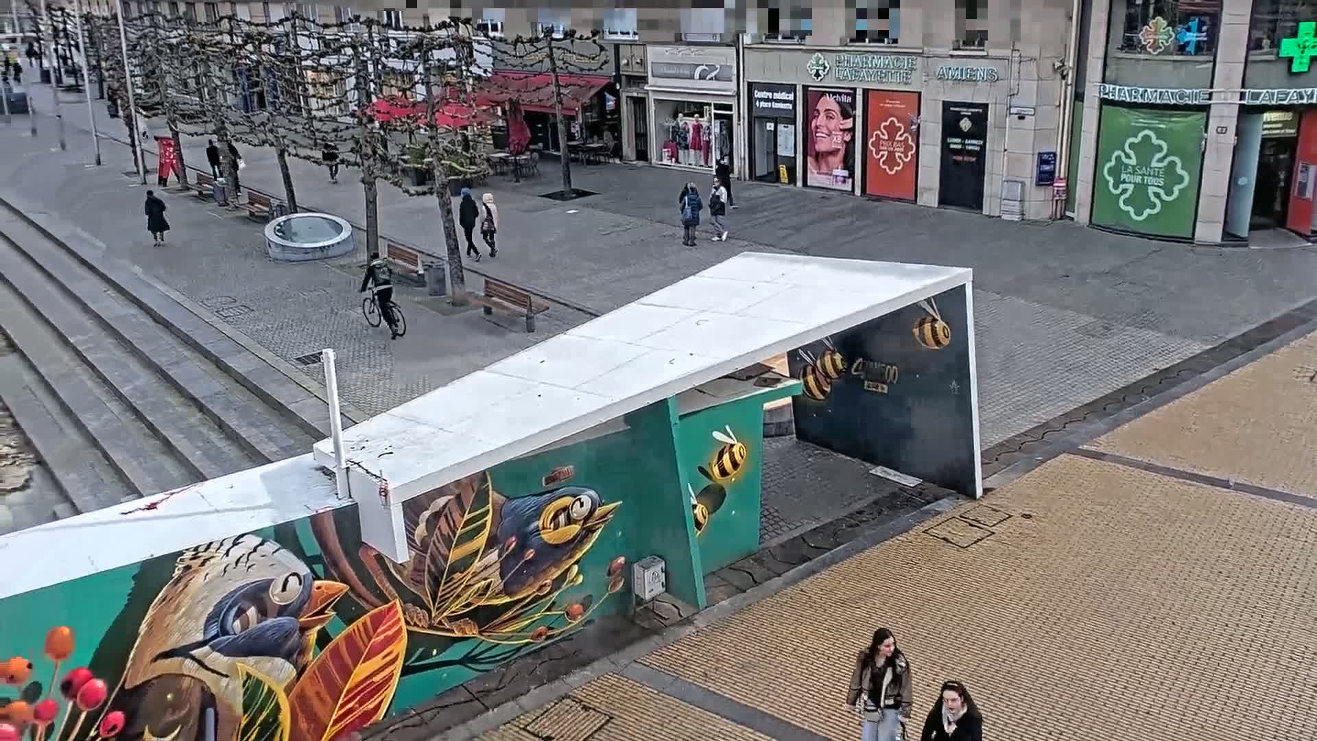 A city street scene on a cloudy day features a mural depicting birds and bees on a wall beneath a white, angular structure, with several pedestrians walking and a cyclist riding past shops and trees.