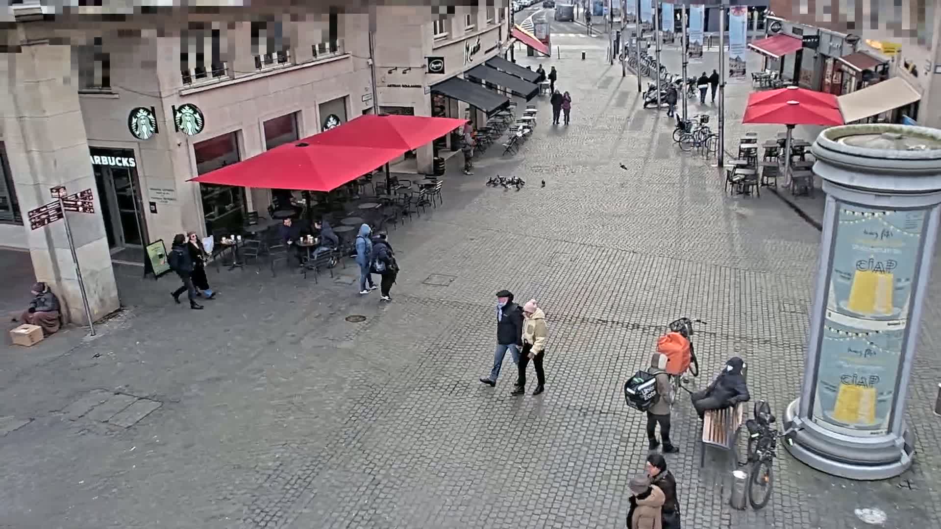 A cobblestone plaza with several people walking, sitting at outdoor cafes, and a few bicycles parked, under an overcast sky.